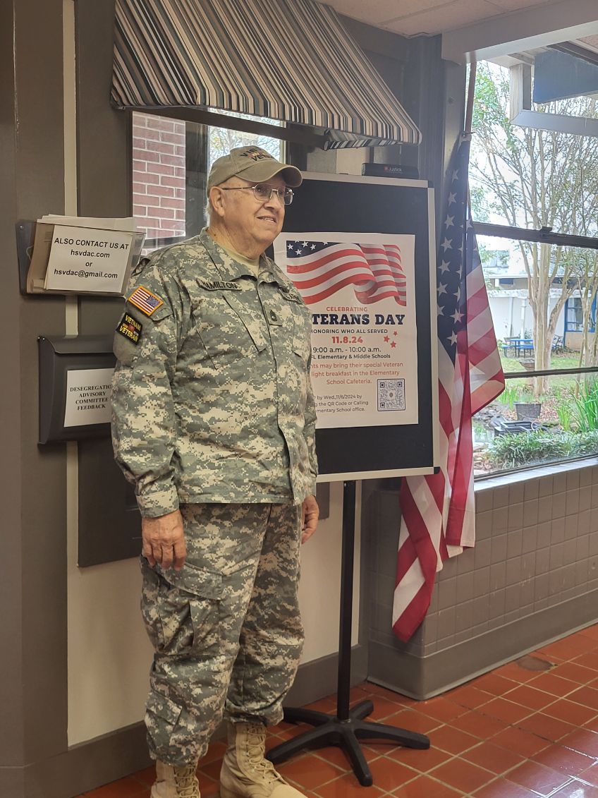 Military Man in Uniform standing next to a Veterans Day sign in the hallway of ASFL Elementary