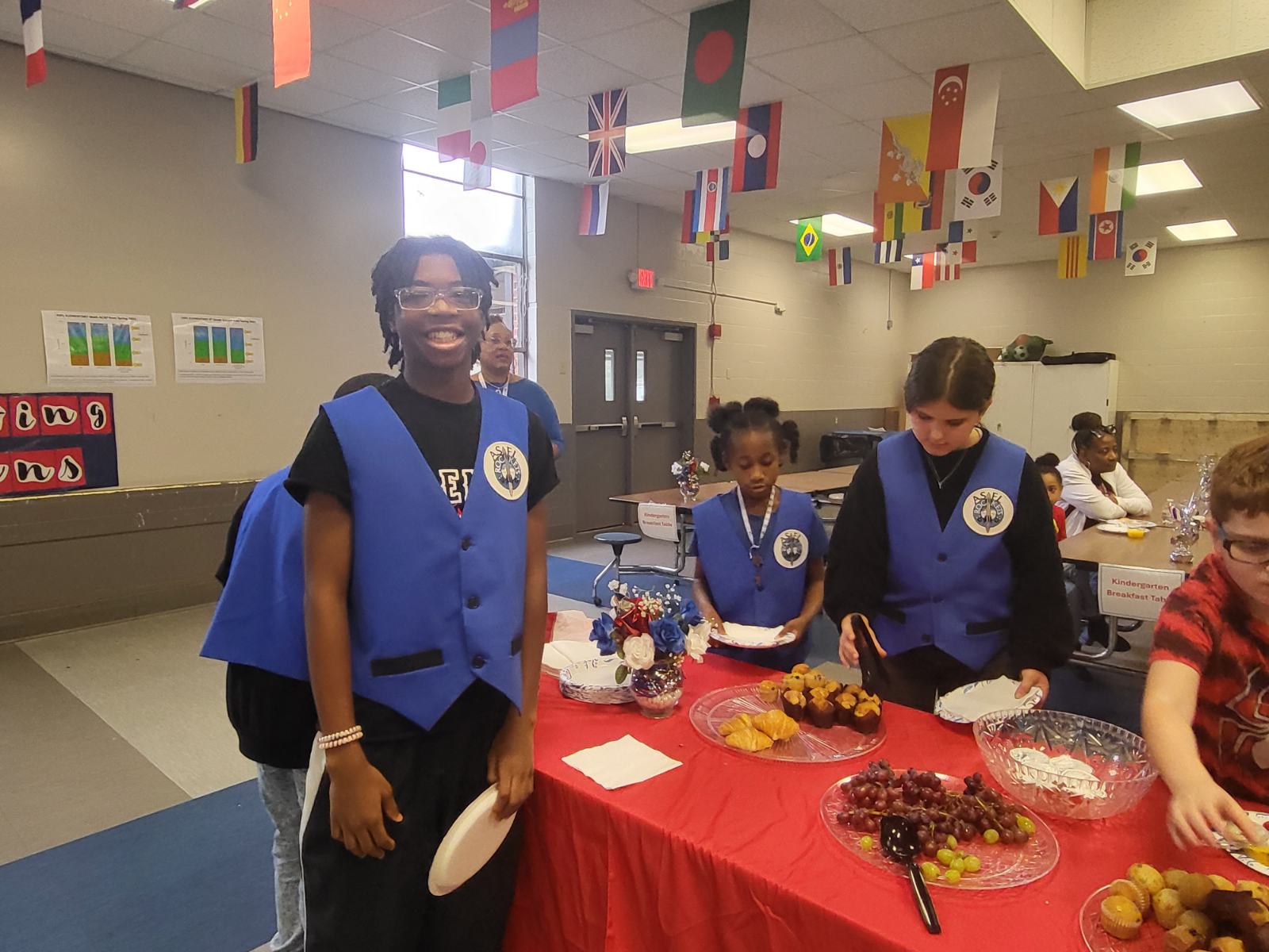 Veterans Day Celebration six students some wearing blue vests serving themselves plates of food. One student standing and smiling at the camera at the academy for science and foreign language elementary