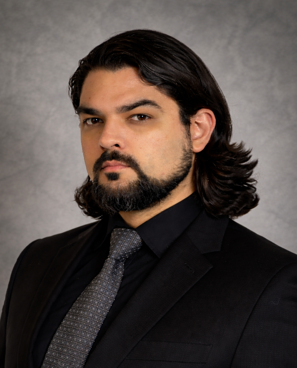 Person in formal attire, wearing a black coat and tie, with long hair and a beard.