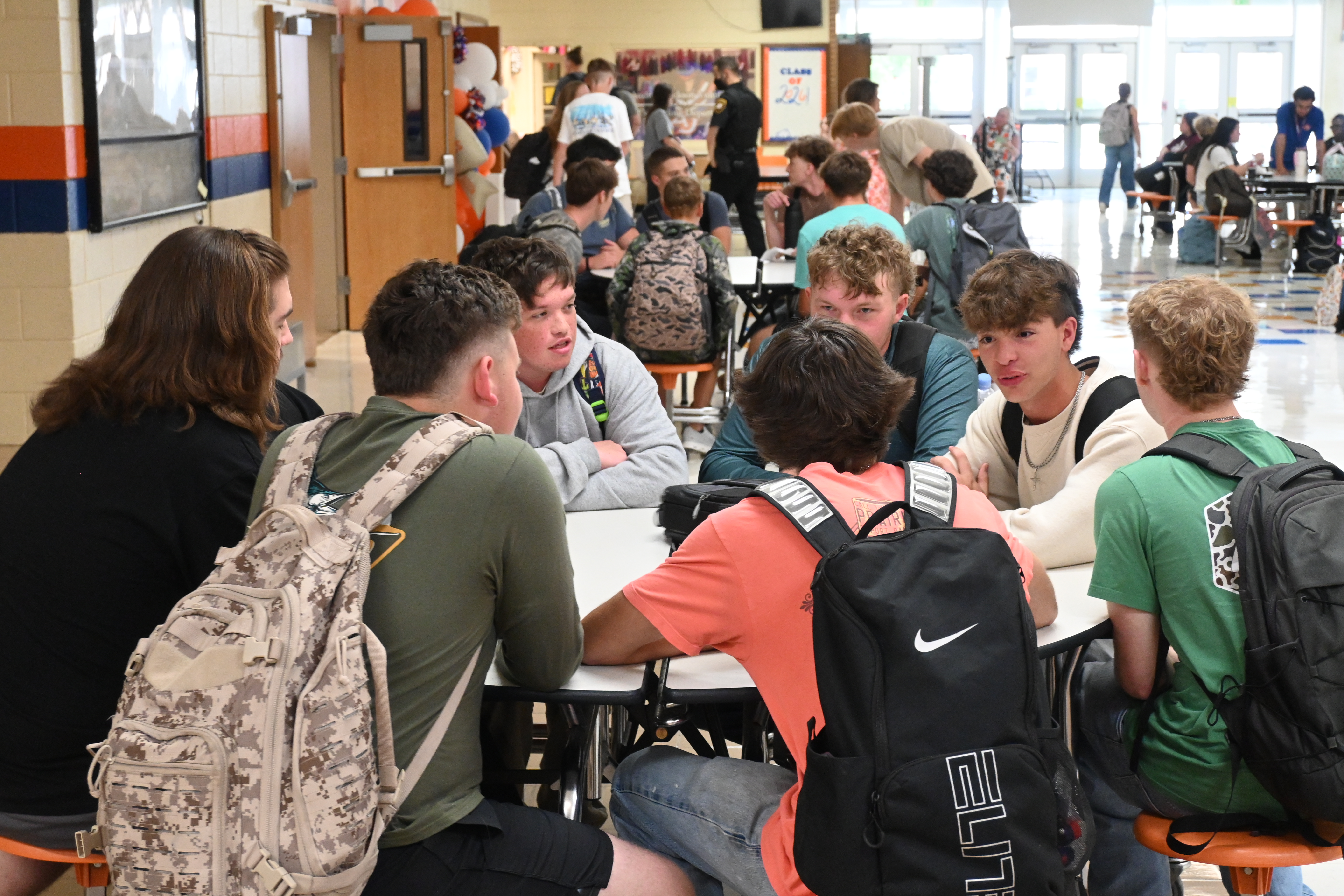 Students wearing backpacks seated around a table inside a school hallway, engaging in a conversation.