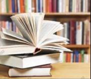 A close-up of a pile of books on a wooden table in a library. The book on the top of the stack is fanned open.