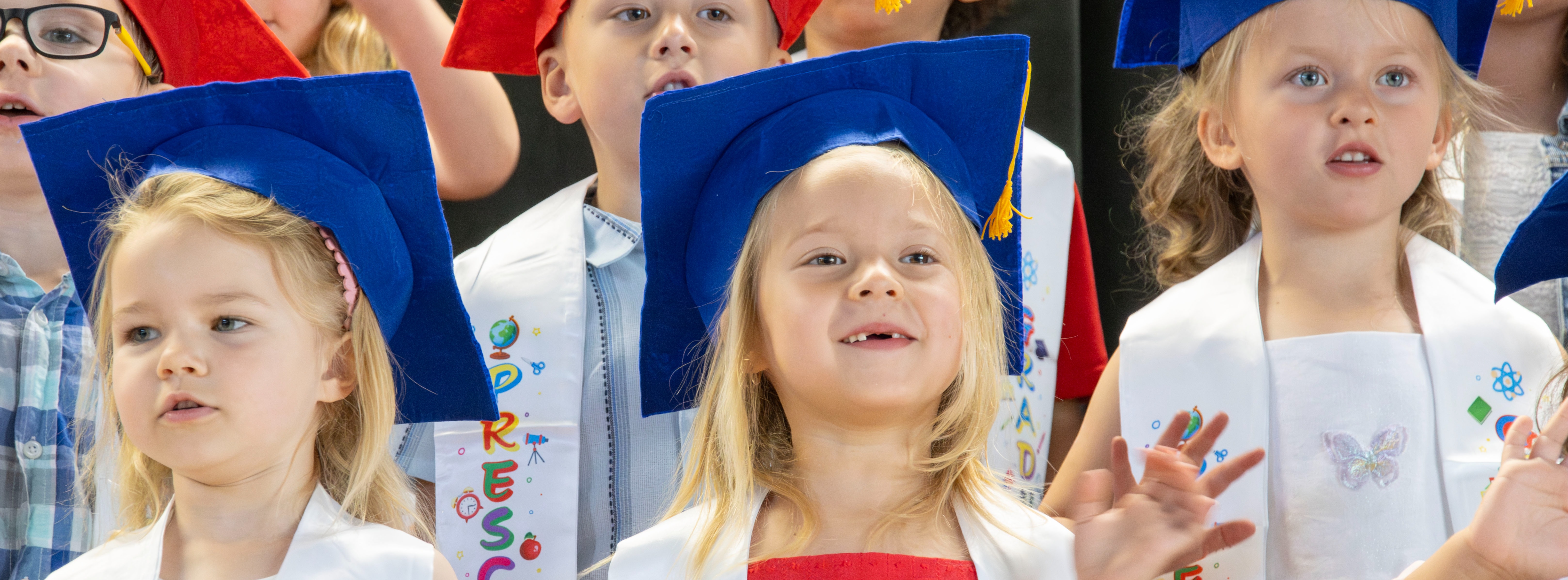 Preschool students wearing red and blue graduation caps and white stoles decorated with colorful school-themed designs participate in a preschool graduation ceremony. Three students in the front row are pictured smiling and gesturing during the ceremony.