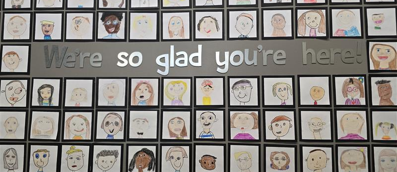 An art installation on a wall with small, square drawings of student faces surround the words “We’re so glad you’re here!” 