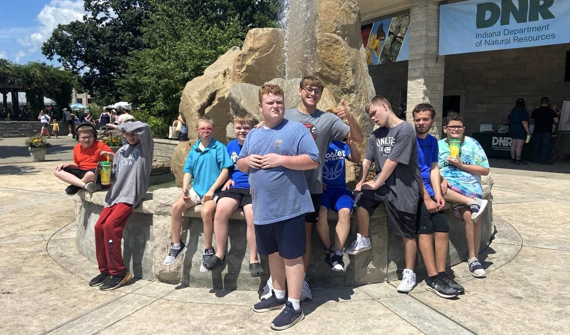 A bright, sunny scene unfolds on a circular plaza in front of a public building marked with a “DNR” sign. Center stage is a large, pale stone fountain or sculptural feature shaped like a four-legged animal; several people are perched on its low ledges and molded contours, using it as a casual group seat. About ten or so individuals—mostly teens and young adults—cluster together in relaxed poses: some sitting, some standing, a few leaning casually against the sculpture.  Clothing is summery and informal—t-shirts in shades of blue, gray, and green, shorts, sneakers, and a couple of caps and sunglasses. The person nearest the camera stands front-and-center in a light blue shirt and dark shorts, hands loosely clasped, while others form a semi-circle behind and to either side, smiling and looking toward the camera. Trees and patches of shade frame the background, and the overall mood feels energetic and convivial, like a group pausing for a quick photo during an outing.