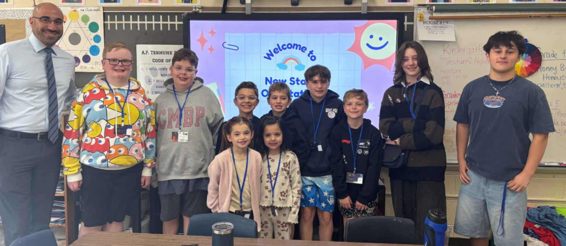 Several students stand next to a teacher, with a screen in the background displaying "Welcome to New York".