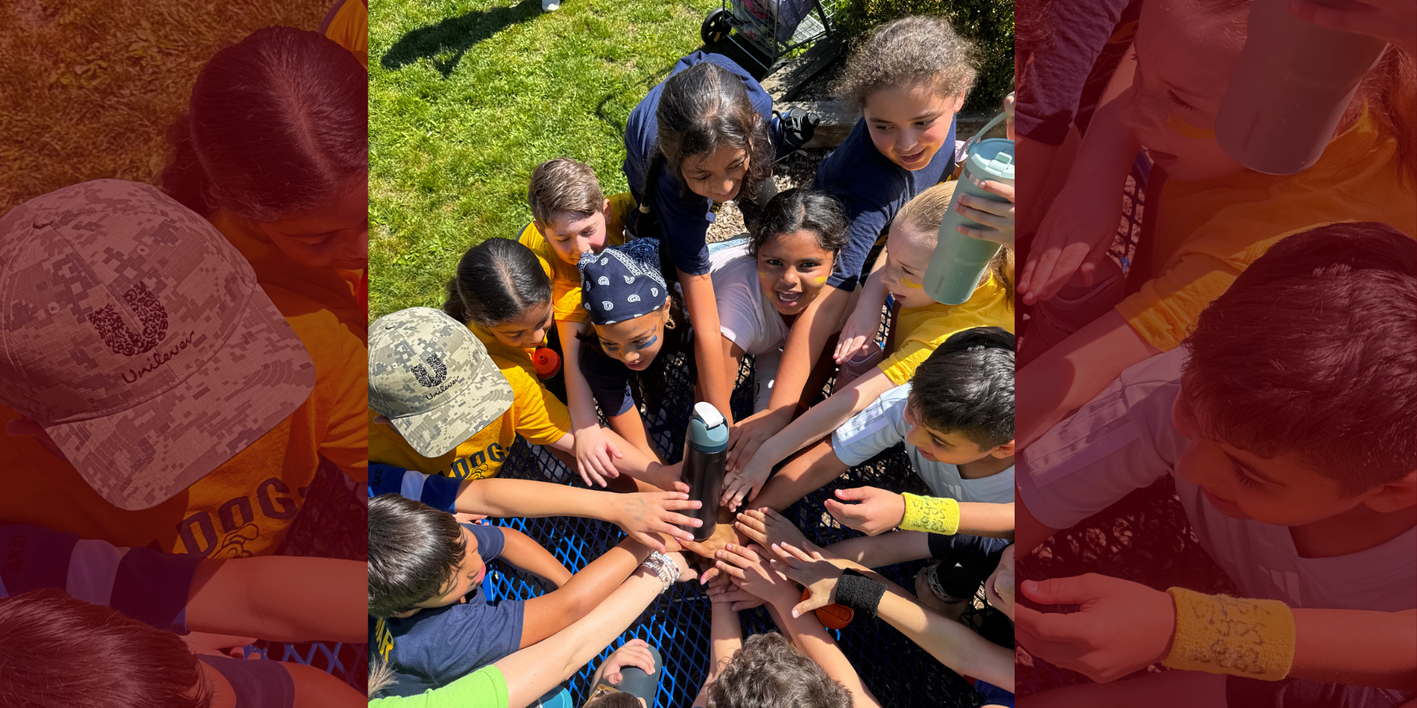 Group of children in orange shirts and hats in a grassy area, hands joined together in a huddle.