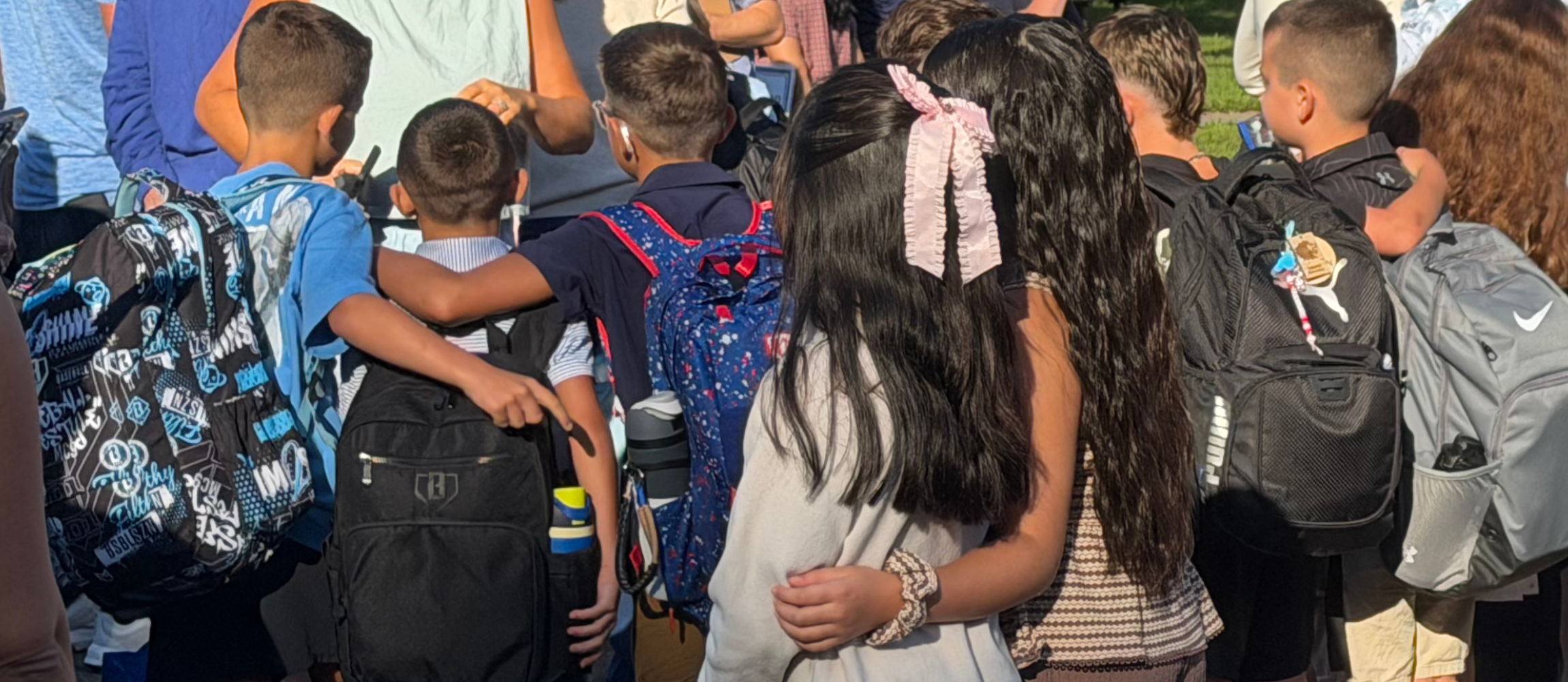 Children stand in a group, wearing backpacks. Some wear bracelets. Background contains other children.