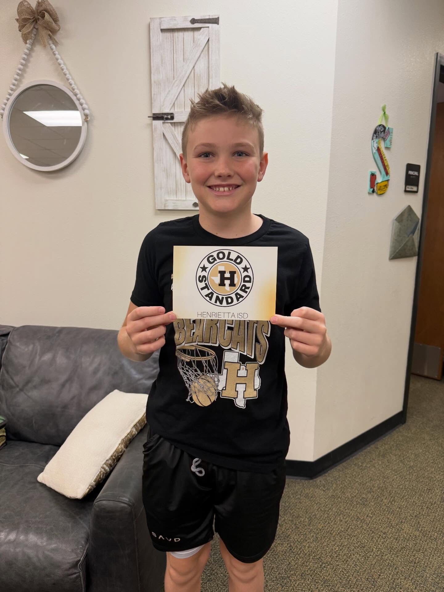 A boy in a black T-shirt and black shorts holds up a certificate in a living room with a gray couch.