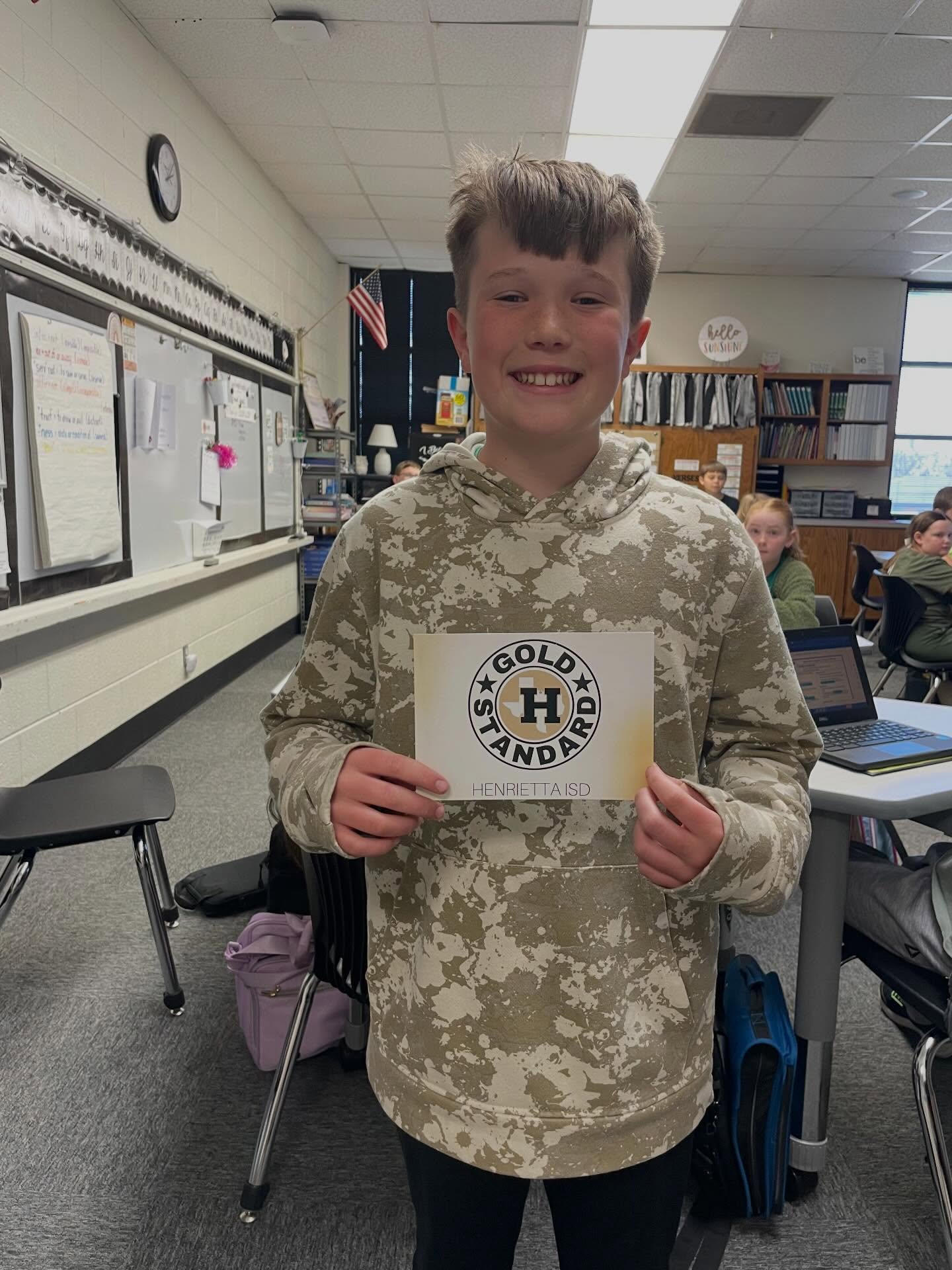 A young boy in a camo hoodie displays a Gold Honor Roll certificate in a classroom. Behind him, shelves, desks, and a clock are visible.