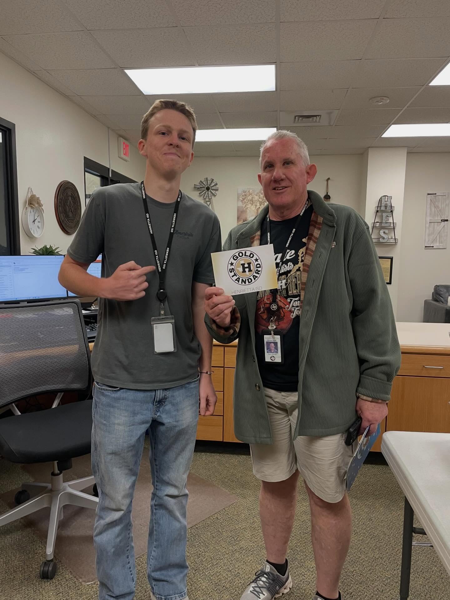 Two men in a room, one holding a card, standing near a desk with a computer and chair.