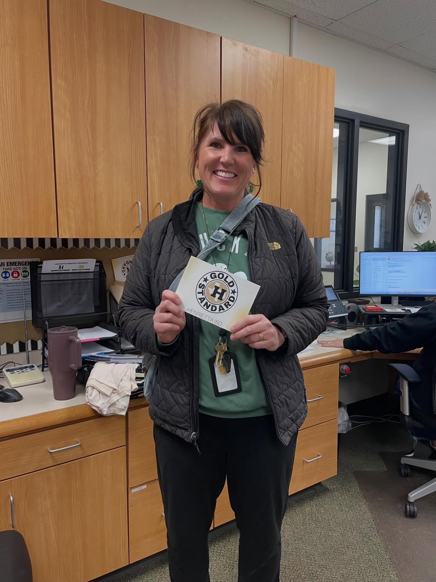 Person standing in an office, holding a paper with text and logo, wearing a green shirt and black pants.