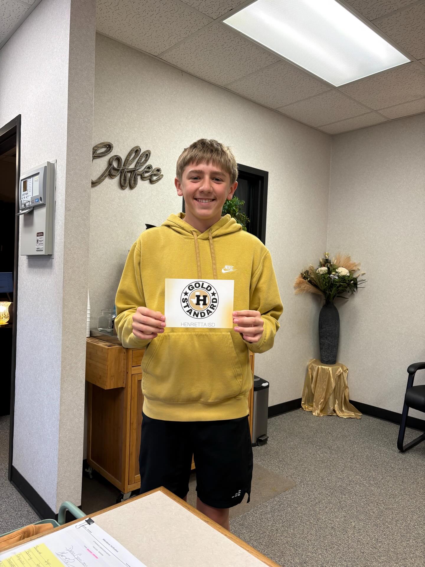 A boy stands in an office, holding a circular sign with text and a logo. Behind him, a wall has "Coffee" written on it.