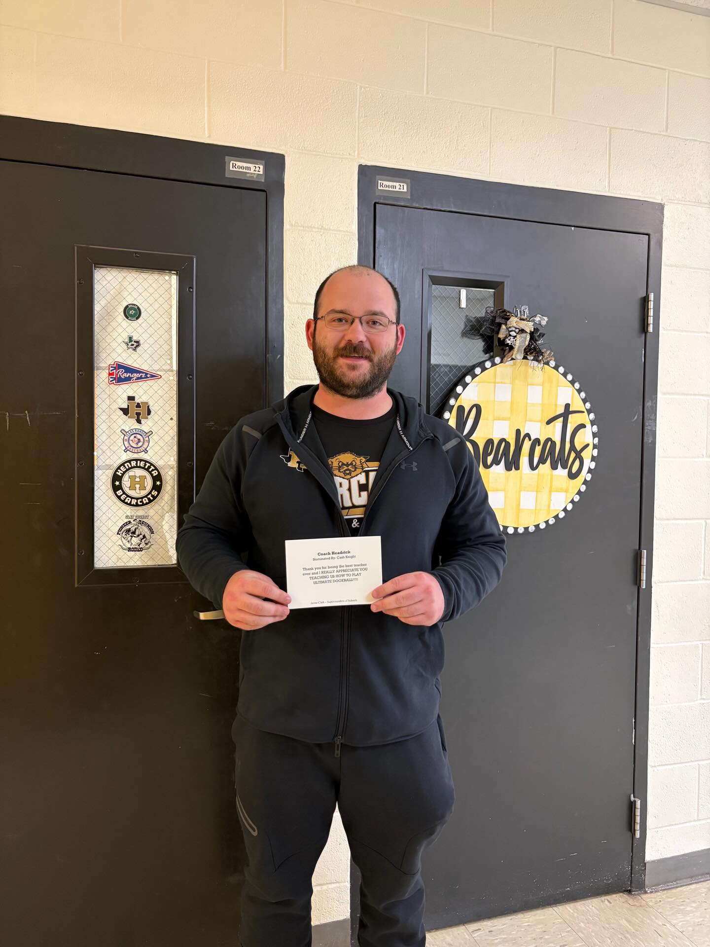 Man in glasses holds a certificate, standing in front of two black doors. One has a glass window with a decorative wreath.