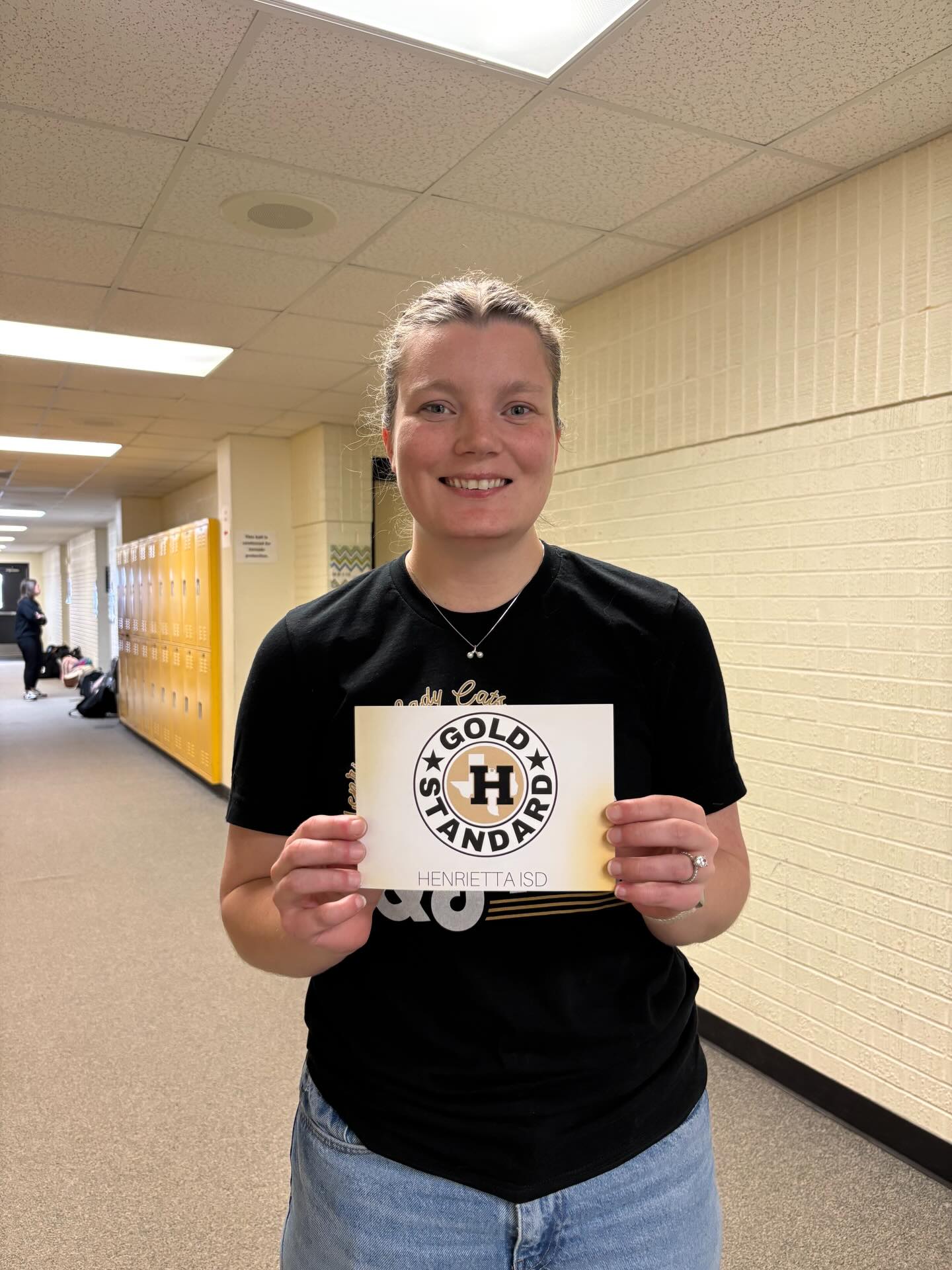 A woman stands in a hallway, holding a yellow sign that reads "Gold Standard". She wears a black shirt and blue jeans.