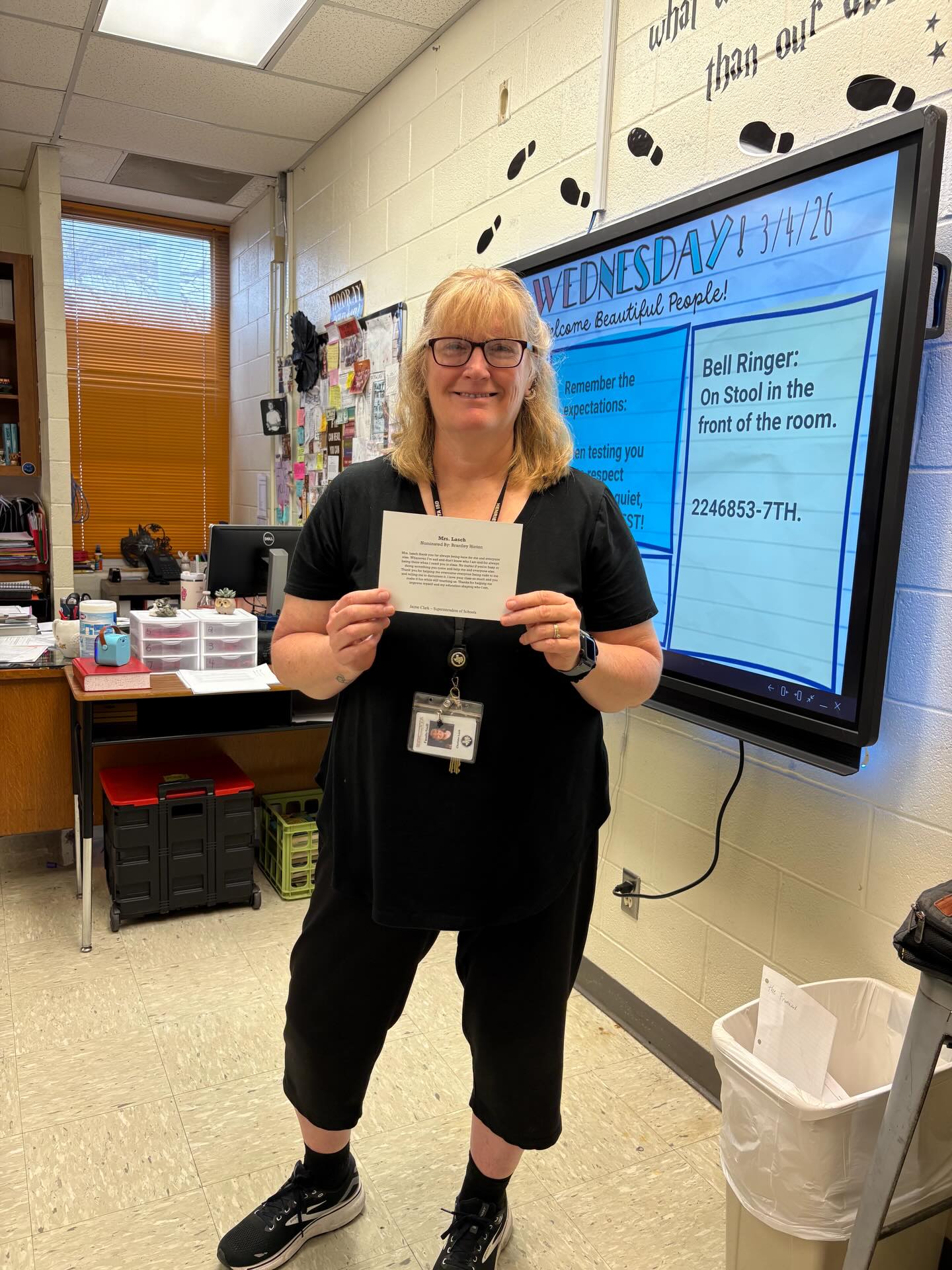 A woman in a black shirt and glasses holds a paper in a classroom. A whiteboard and desk are behind her.