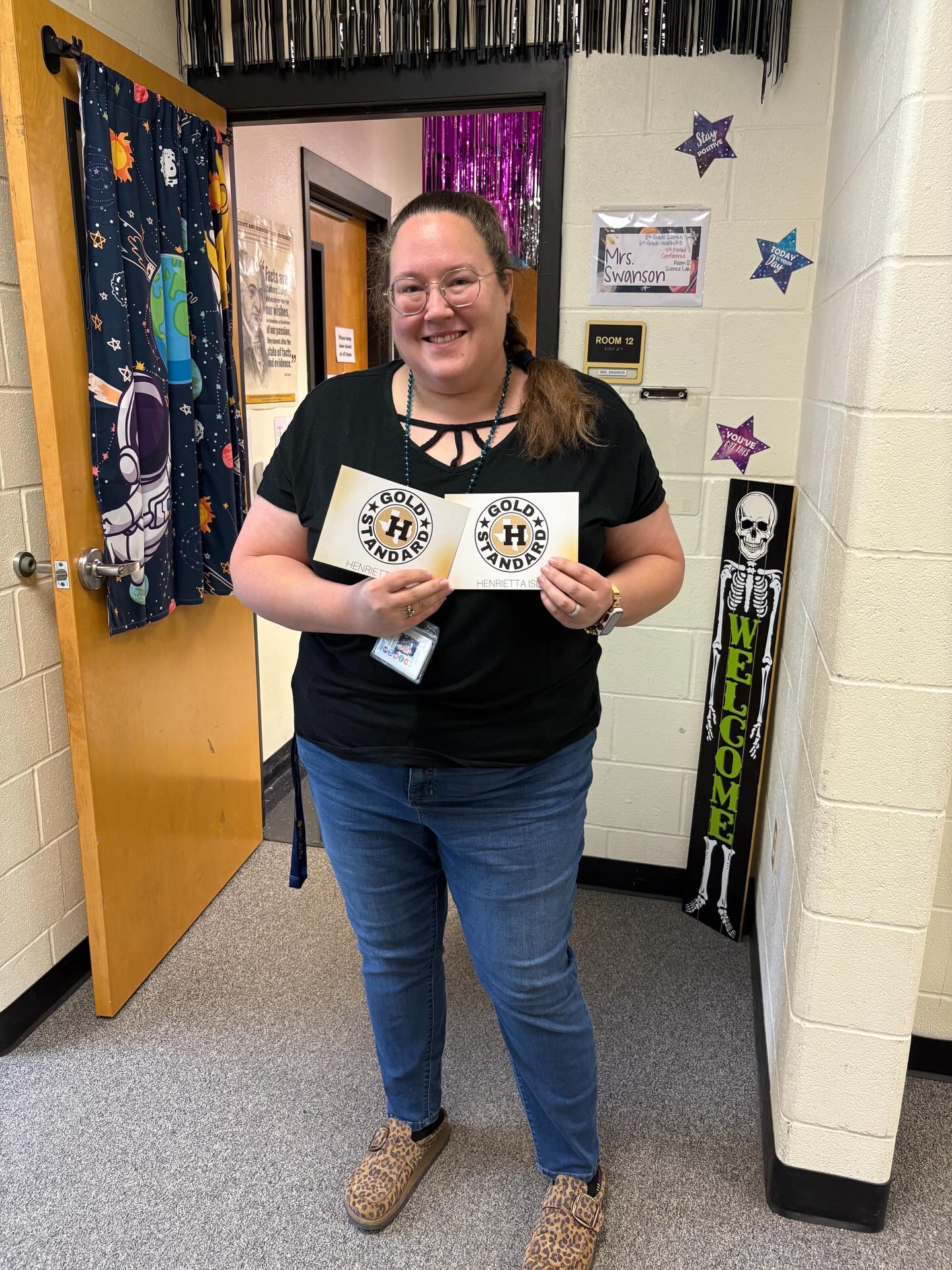 Woman in black shirt holding two white cards, standing in a classroom hallway with a door and decorations.