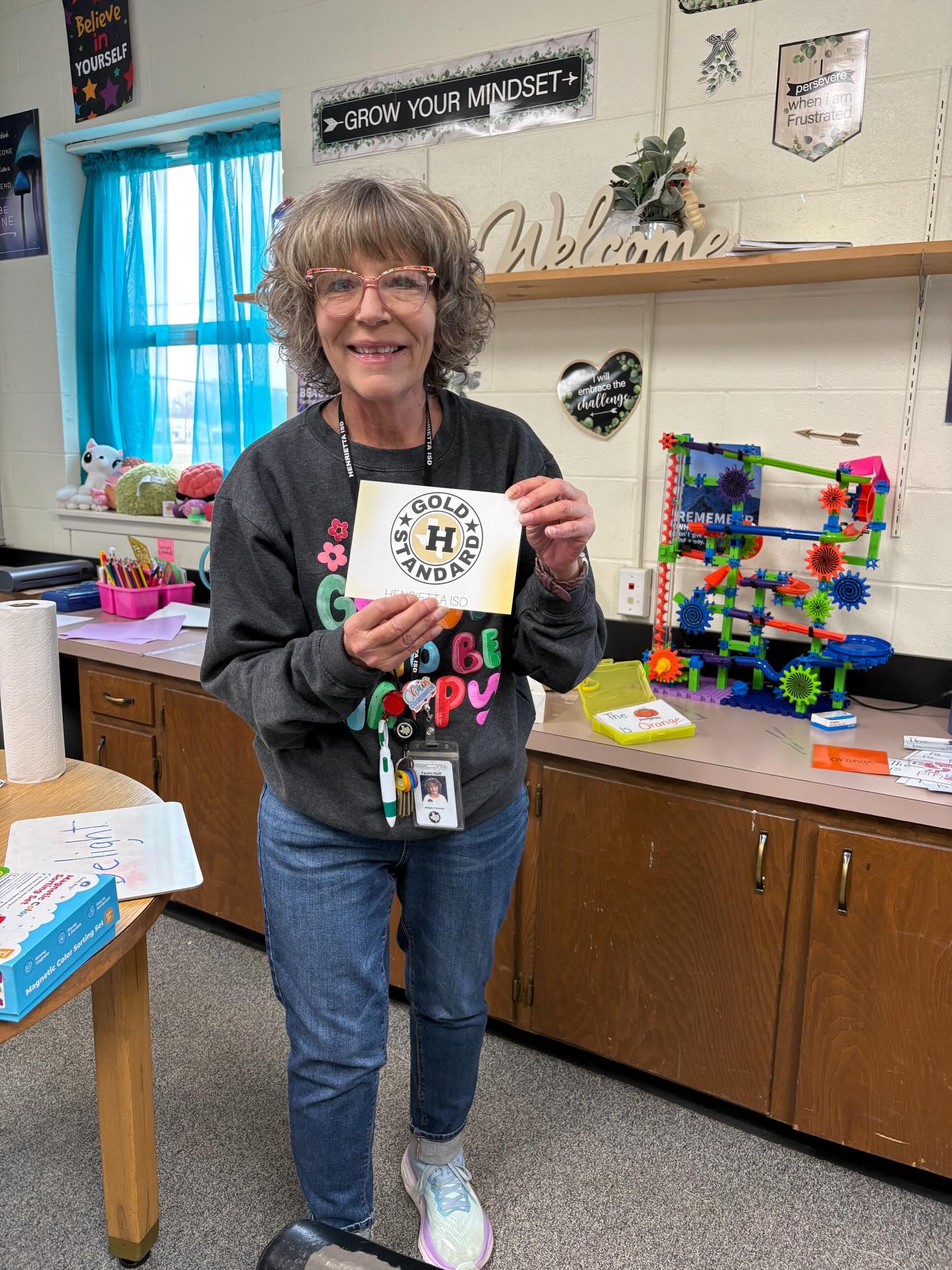 A person holds a certificate in a classroom with a table, shelves, and a window with a blue curtain.
