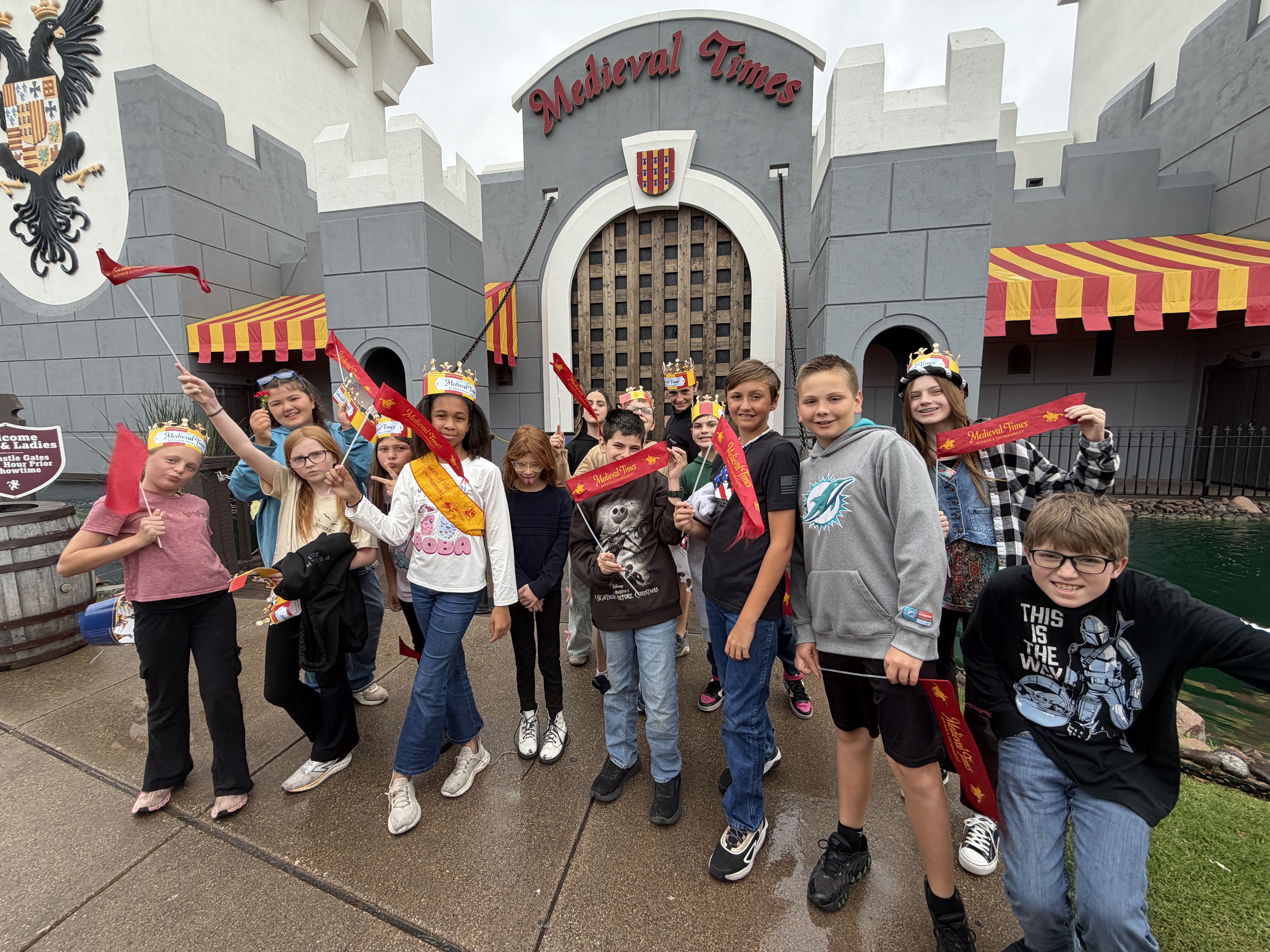 A group of children, some wearing crowns and holding flags, poses for a photo in front of a medieval-themed building.