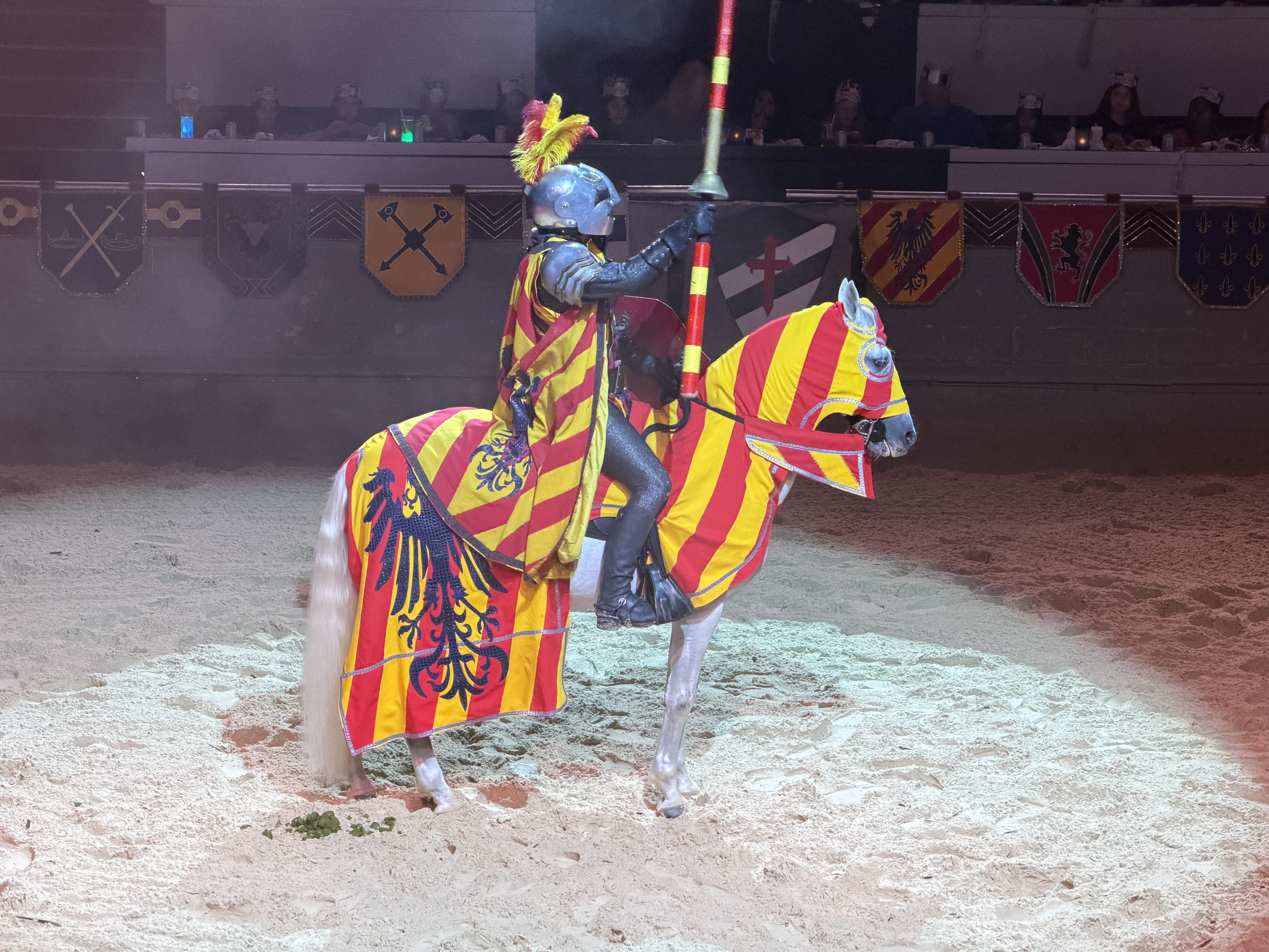 Person in knight's armor and helmet riding a horse with a red and yellow cape.