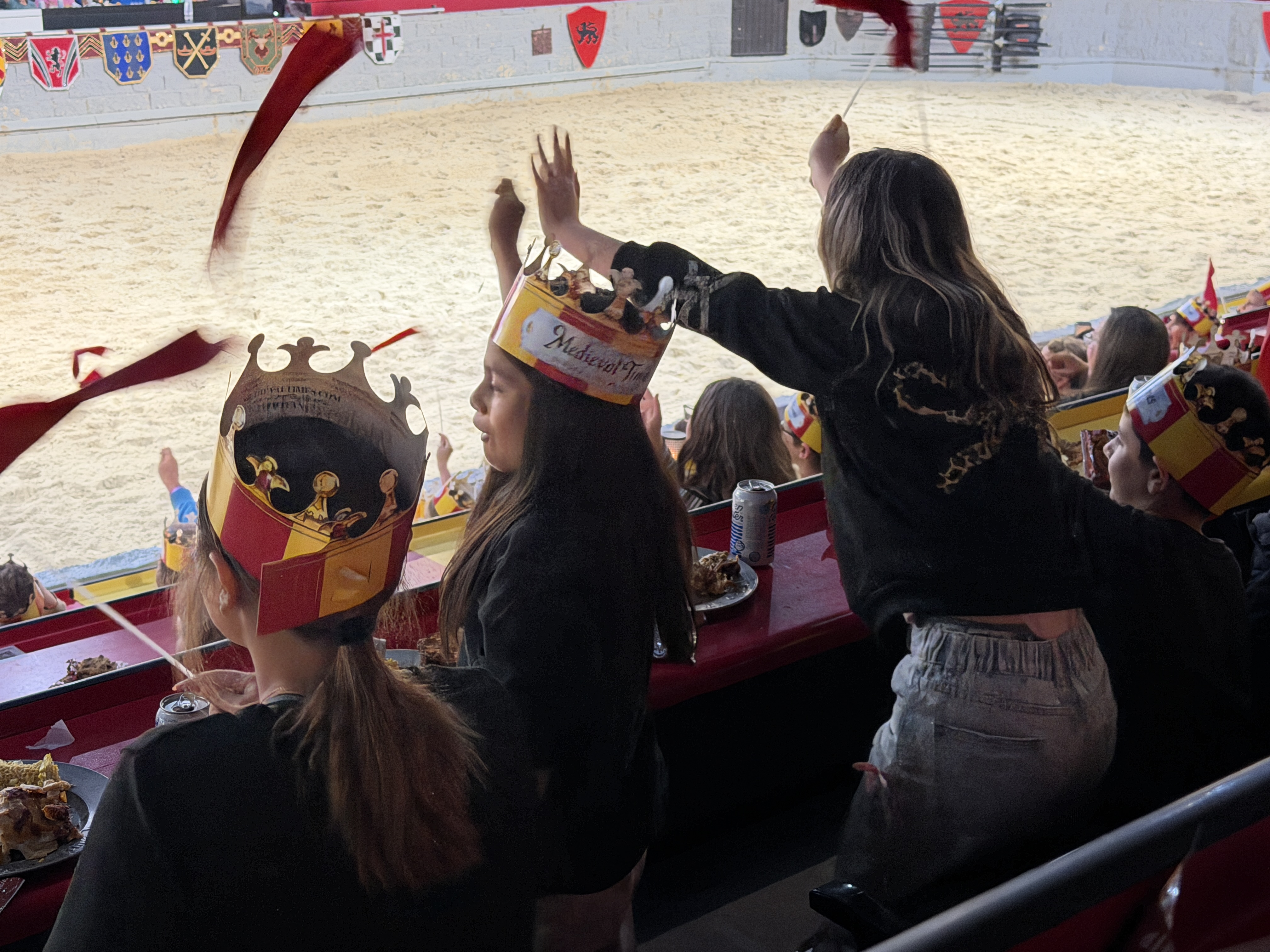 Spectators wear crowns as they cheer in a stadium. The audience sits at tables with food.