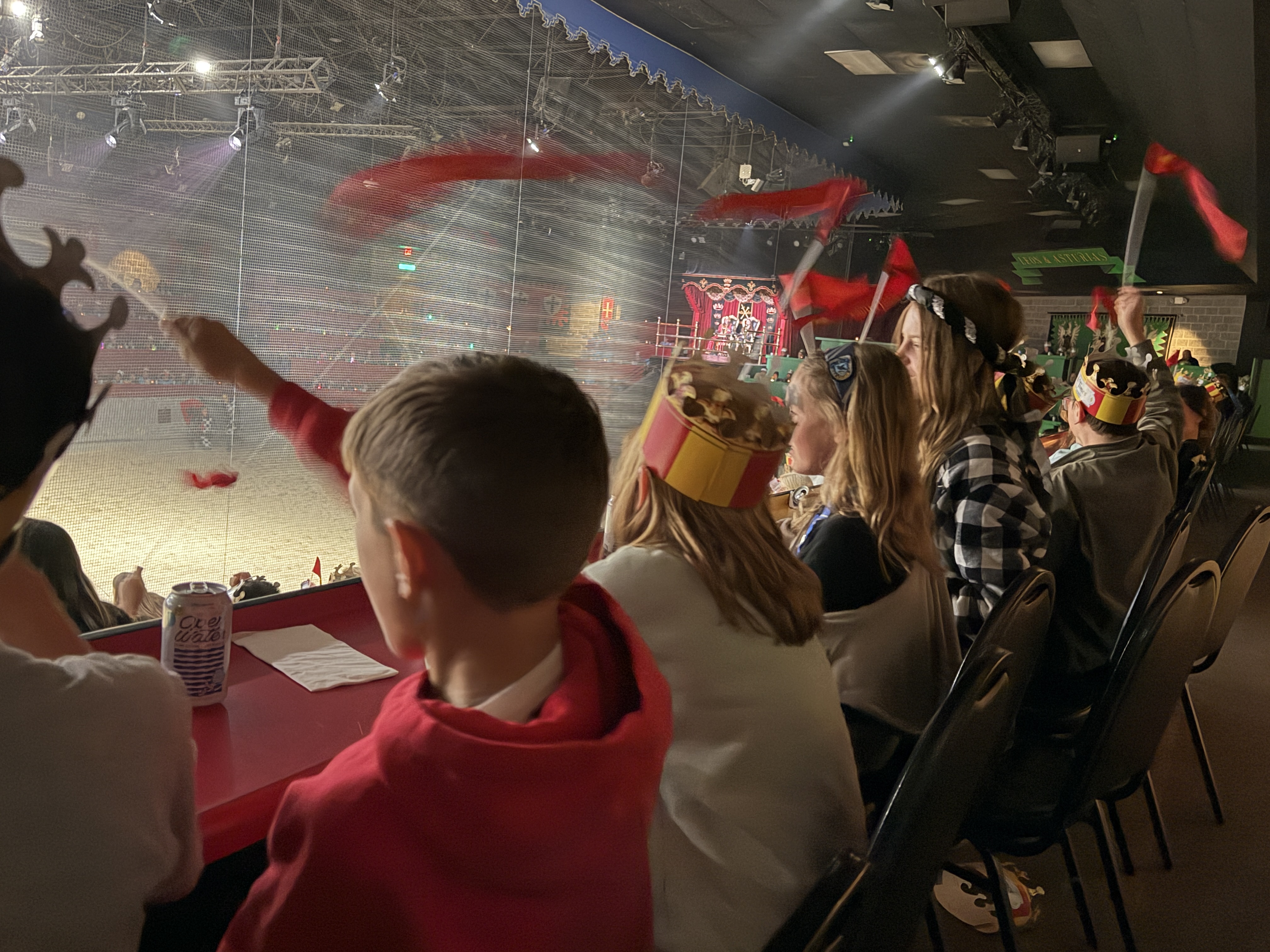 Several kids in crowns sit at a table watching a jousting competition through a large window.