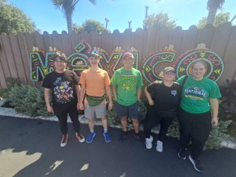 Five individuals stand together in front of a wooden fence with colorful graffiti. One wears a green shirt with text.