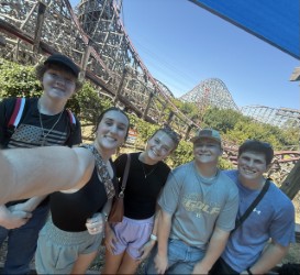 Five people stand before a roller coaster. They smile and pose for a photograph.