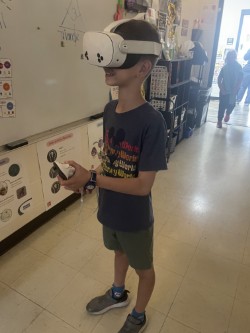 A child wearing a white virtual reality headset, holding a controller in a classroom with white walls and tiled floors.