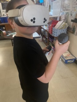 A young boy wears a white virtual reality headset and holds two controllers. Shelves and items are in the background.