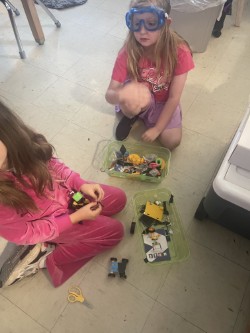 Two young girls in pink outfits are on the floor, using scissors and building with plastic toys.