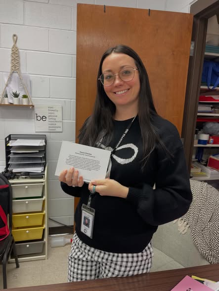 Woman in glasses holding certificate. Behind her, a door, drawers, a shelf with items, and a white wall.