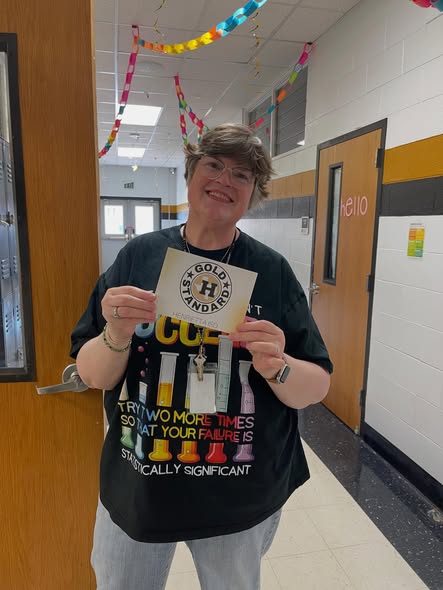 Person with a black tee, a necklace, and a ring, holding a yellow card, in a hallway with doors and decorations.