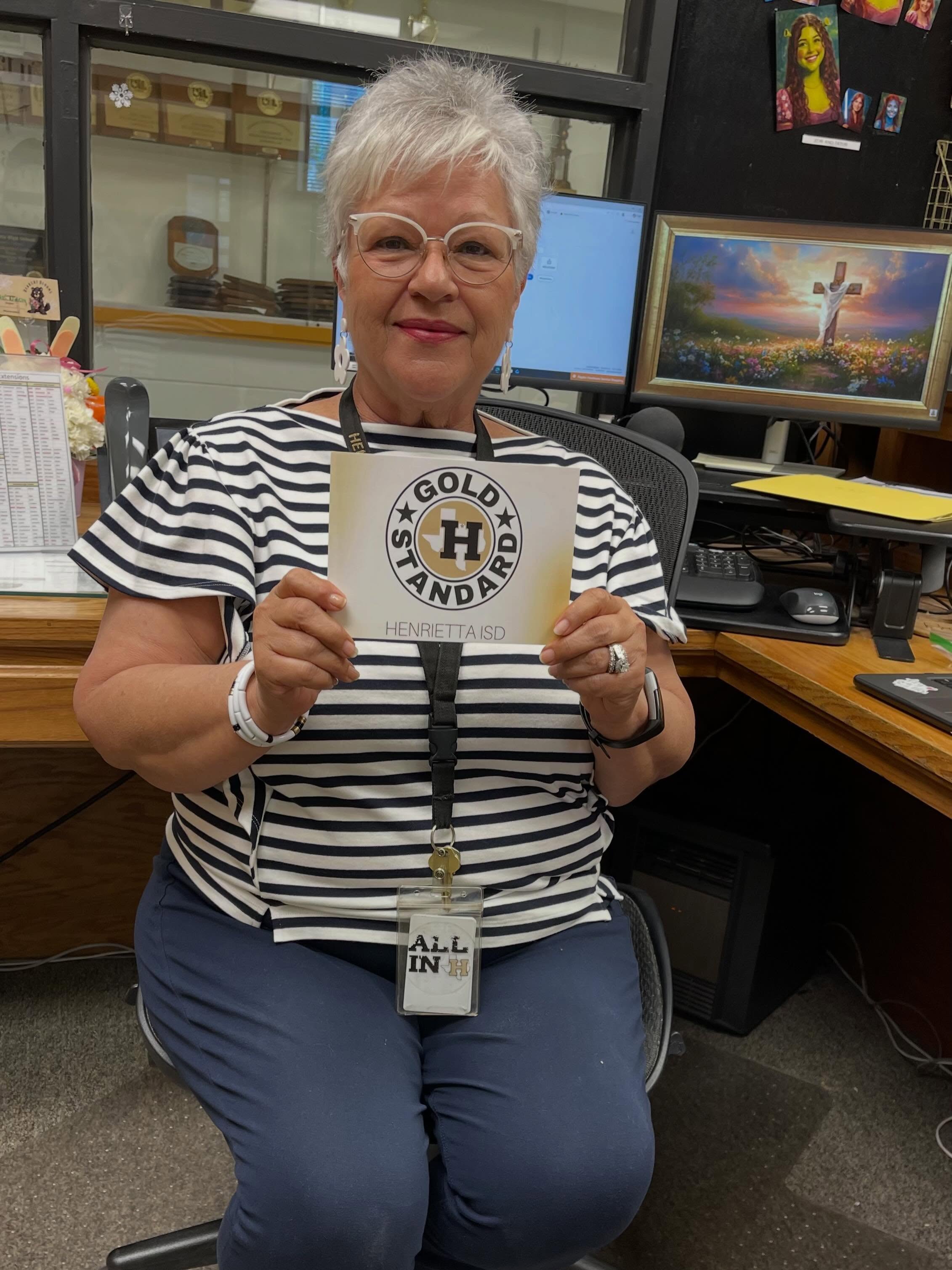 Woman in a black and white striped shirt holds a card that says "GOLD STANDARD" at an office desk.