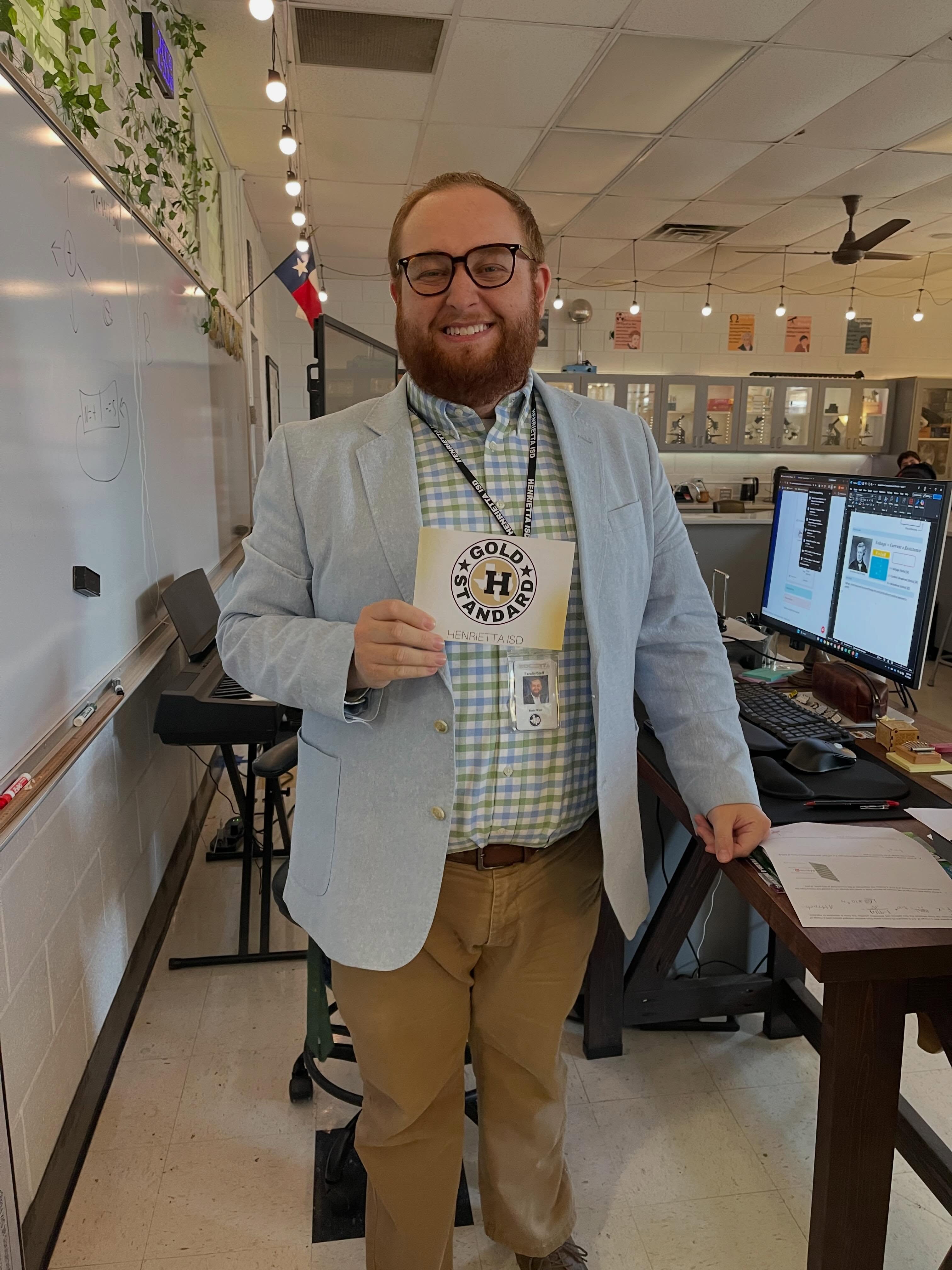 A man in a light blue blazer and glasses stands in a classroom holding a yellow card.