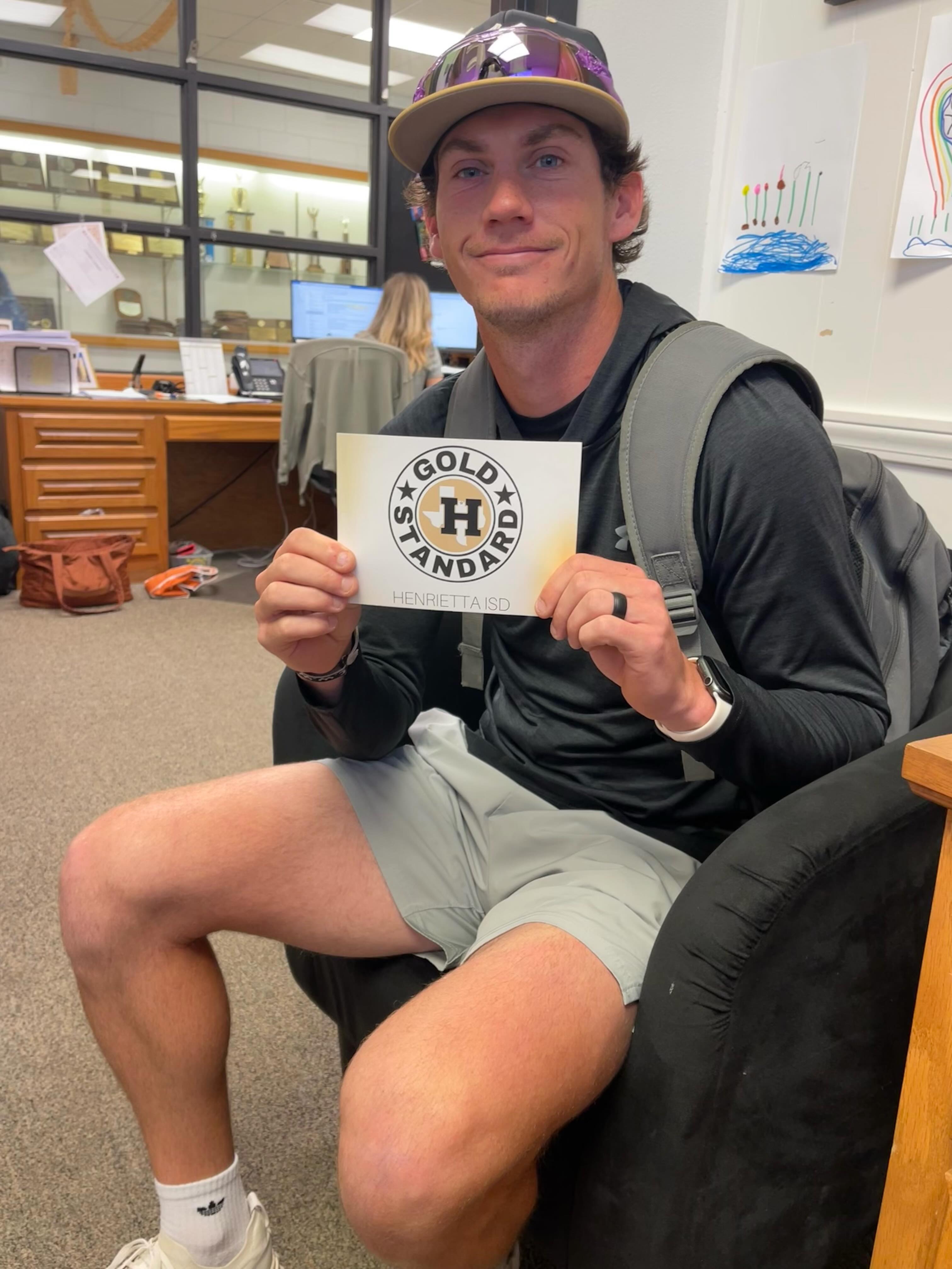 Man in cap and backpack holds sign with text "Gold Standard" while sitting in a chair.