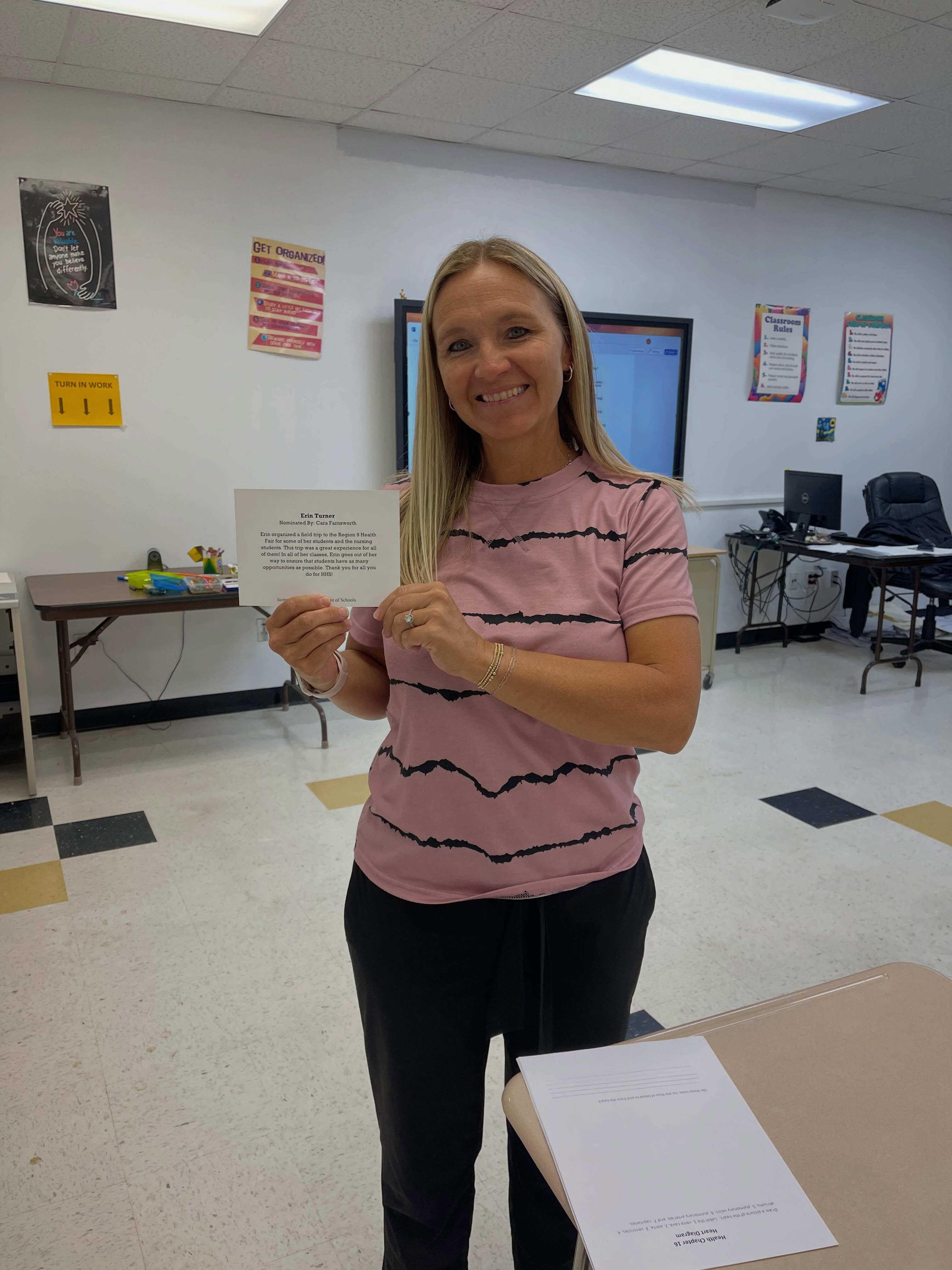 A woman stands in a classroom, holding a certificate, smiling. Behind her, tables and chairs are set up, and a monitor and bulletin board are on the wall.