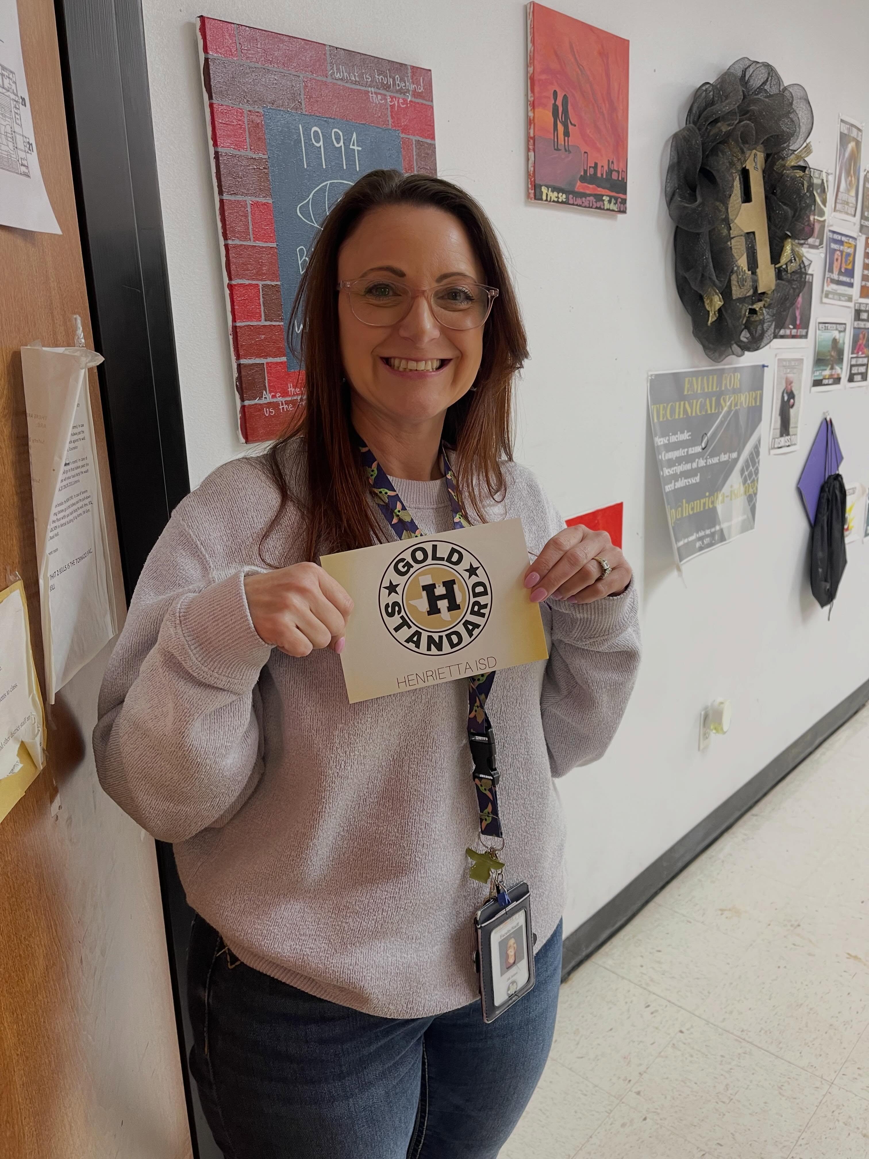 A smiling woman in a white sweatshirt holds a "Gold Standard" certificate in a school hallway.
