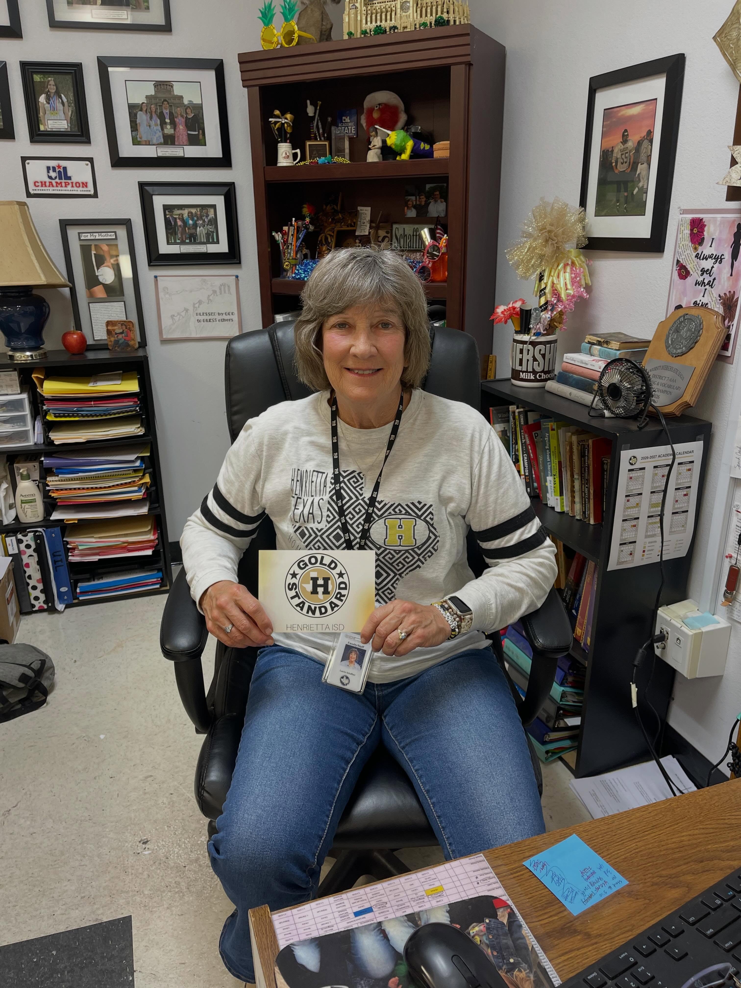 An adult woman holds a card and sits in a chair in front of a desk. Shelves with books and framed photos adorn the background.