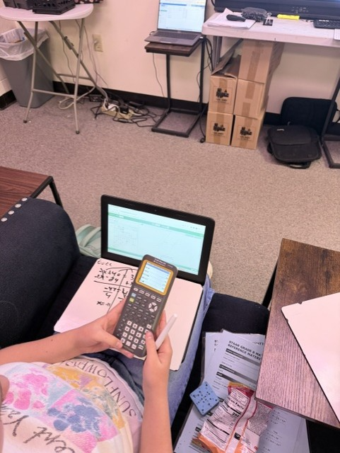 Person sitting on a chair with a calculator, laptop, and papers. A table with a white paper and other objects in the background.