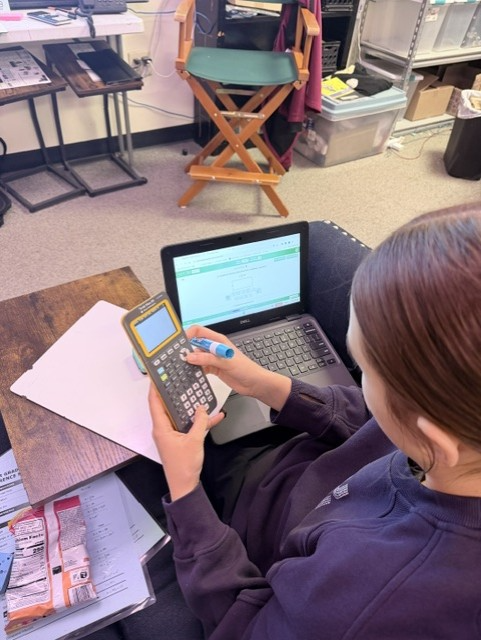 Person using a calculator in a classroom. A laptop, paper, and chair are in the background.