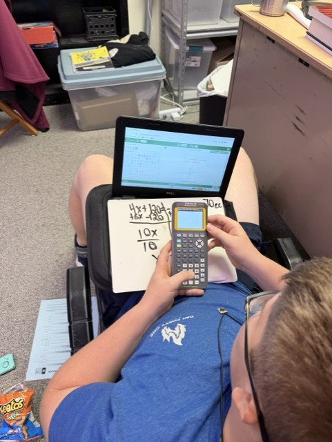 A student uses a calculator and tablet in a classroom. Papers with handwritten and typed math problems are on the desk.