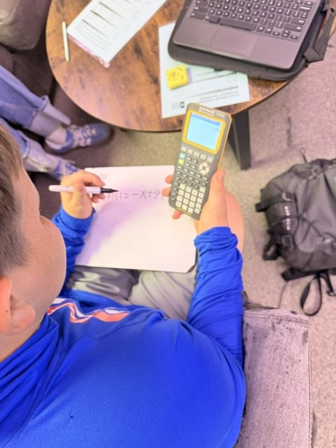 A person uses a calculator, writing on paper. A table has a laptop, papers, and a marker.