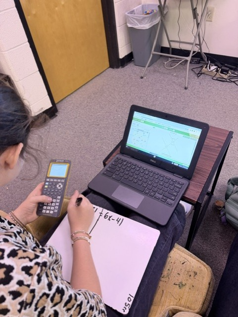 A person sits at a desk, holding a calculator and writing on a notebook. A laptop sits open on the desk.