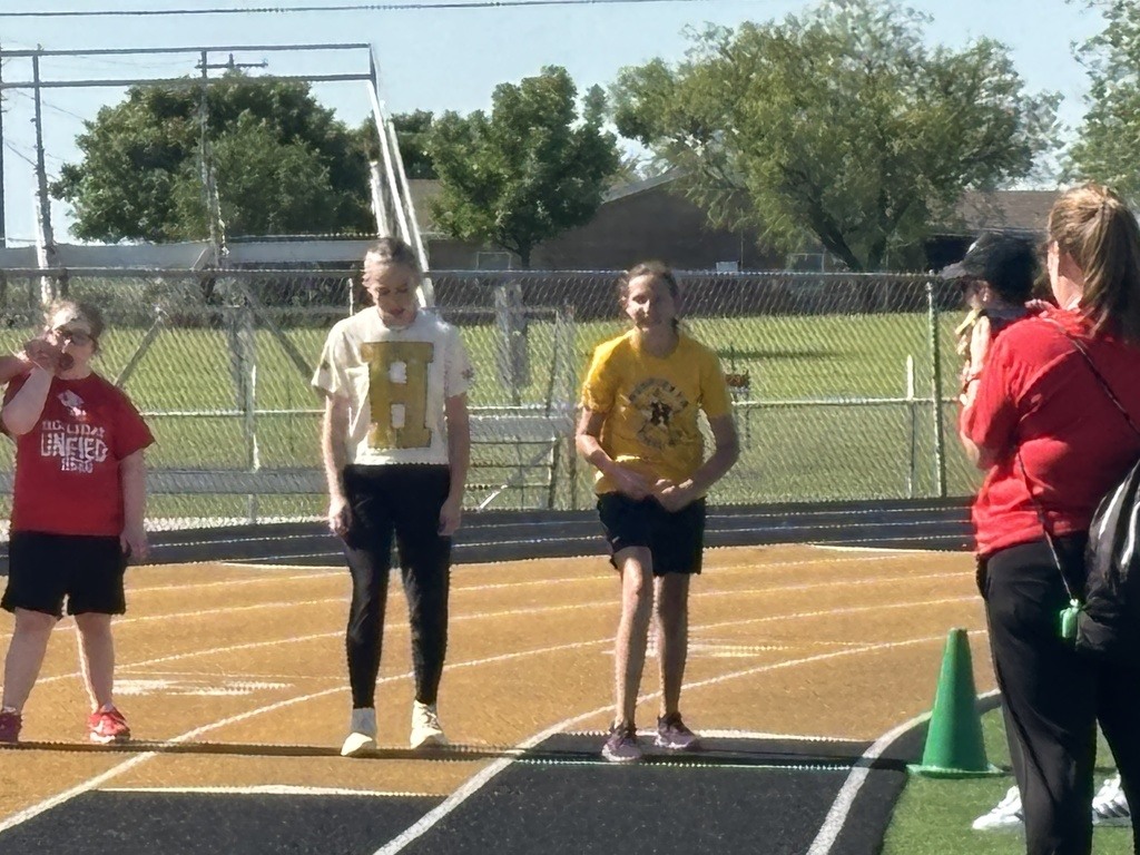 Four people stand on a track, two wearing red shirts. A metal fence and trees surround the track.