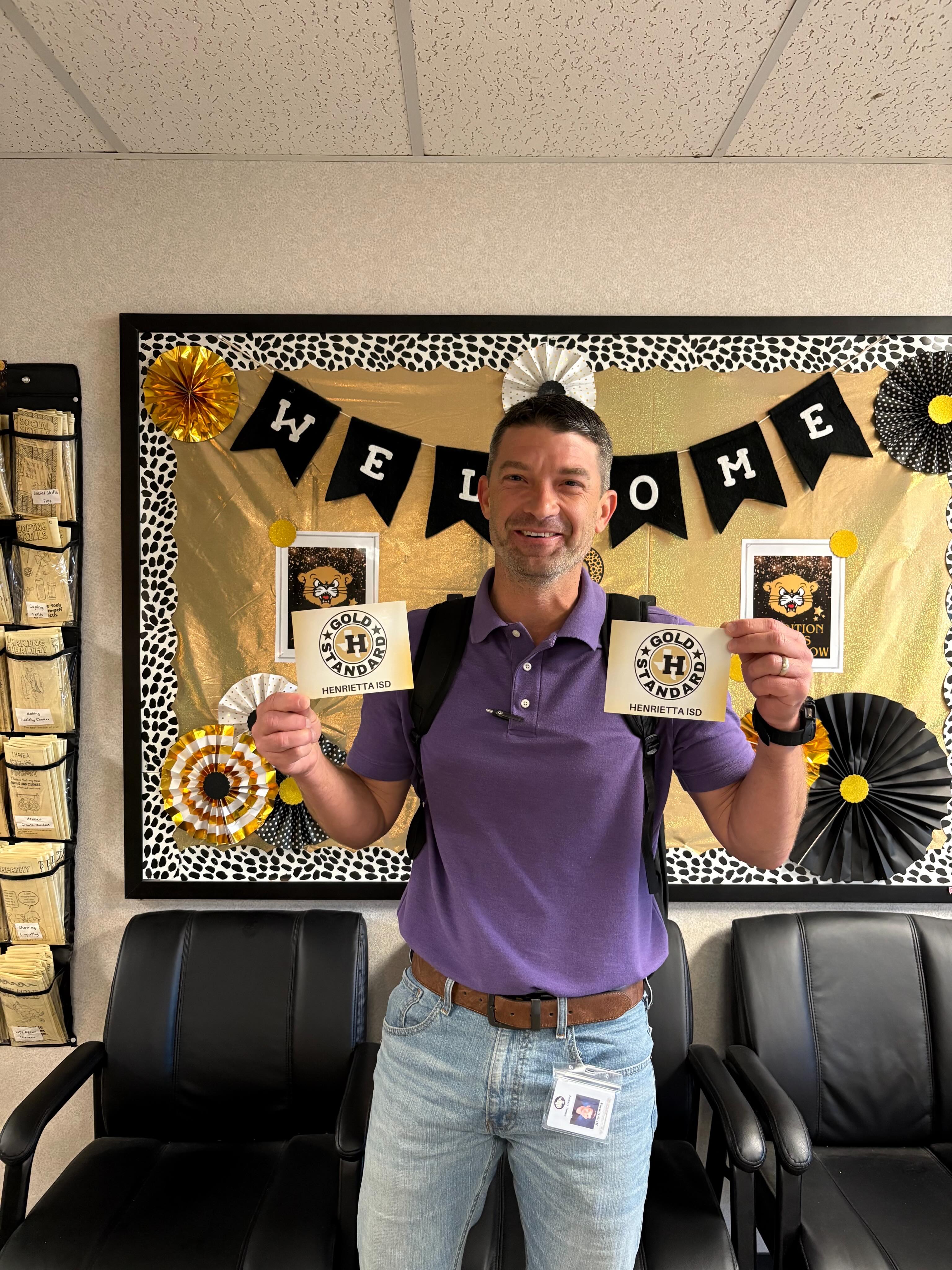 A man in a purple shirt and jeans holds up two cards, standing in front of a decorative banner with "Welcome" on it.
