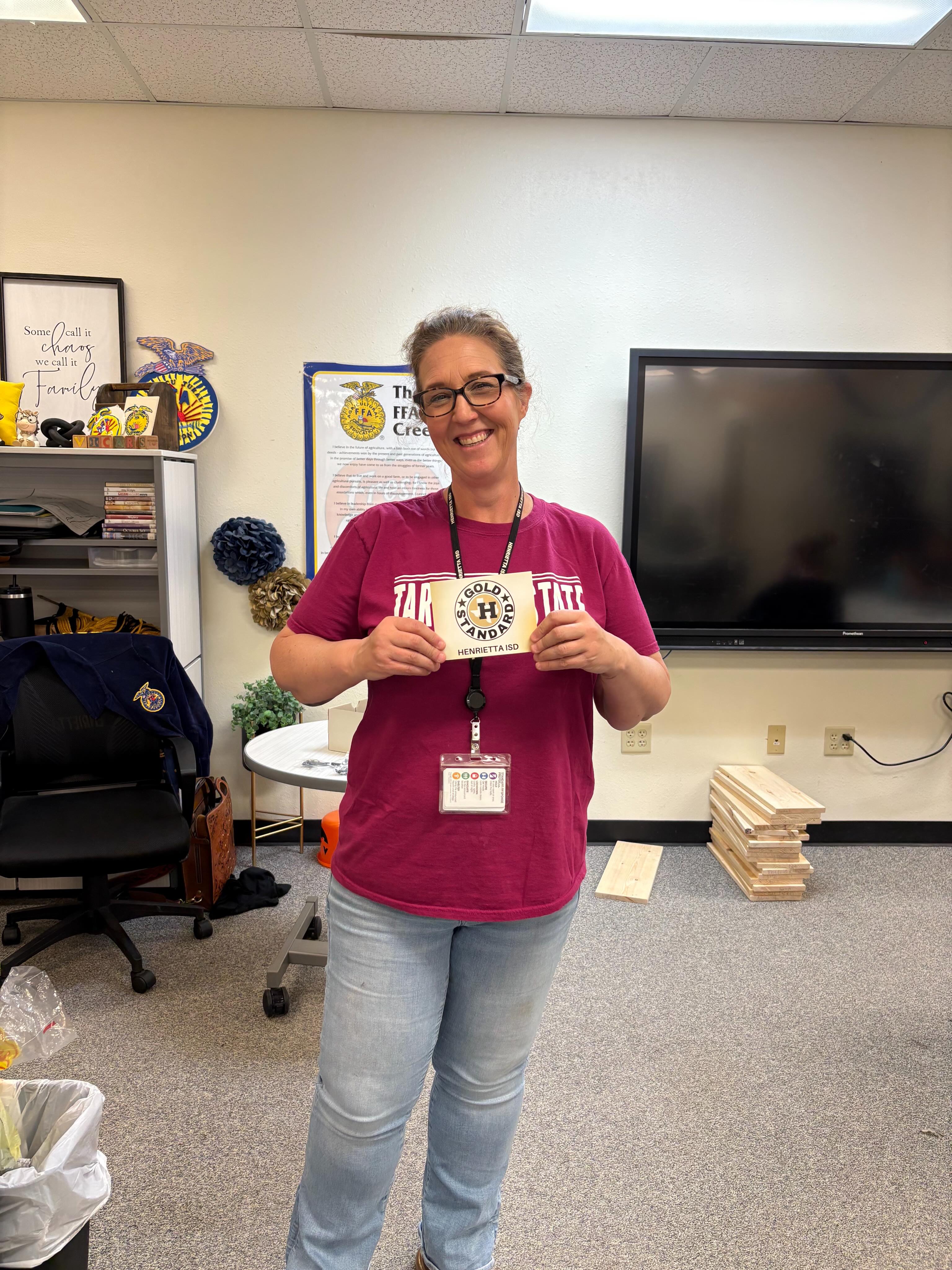 Person in purple shirt holding a card with a badge, standing in an office with a monitor, chair, and desk.