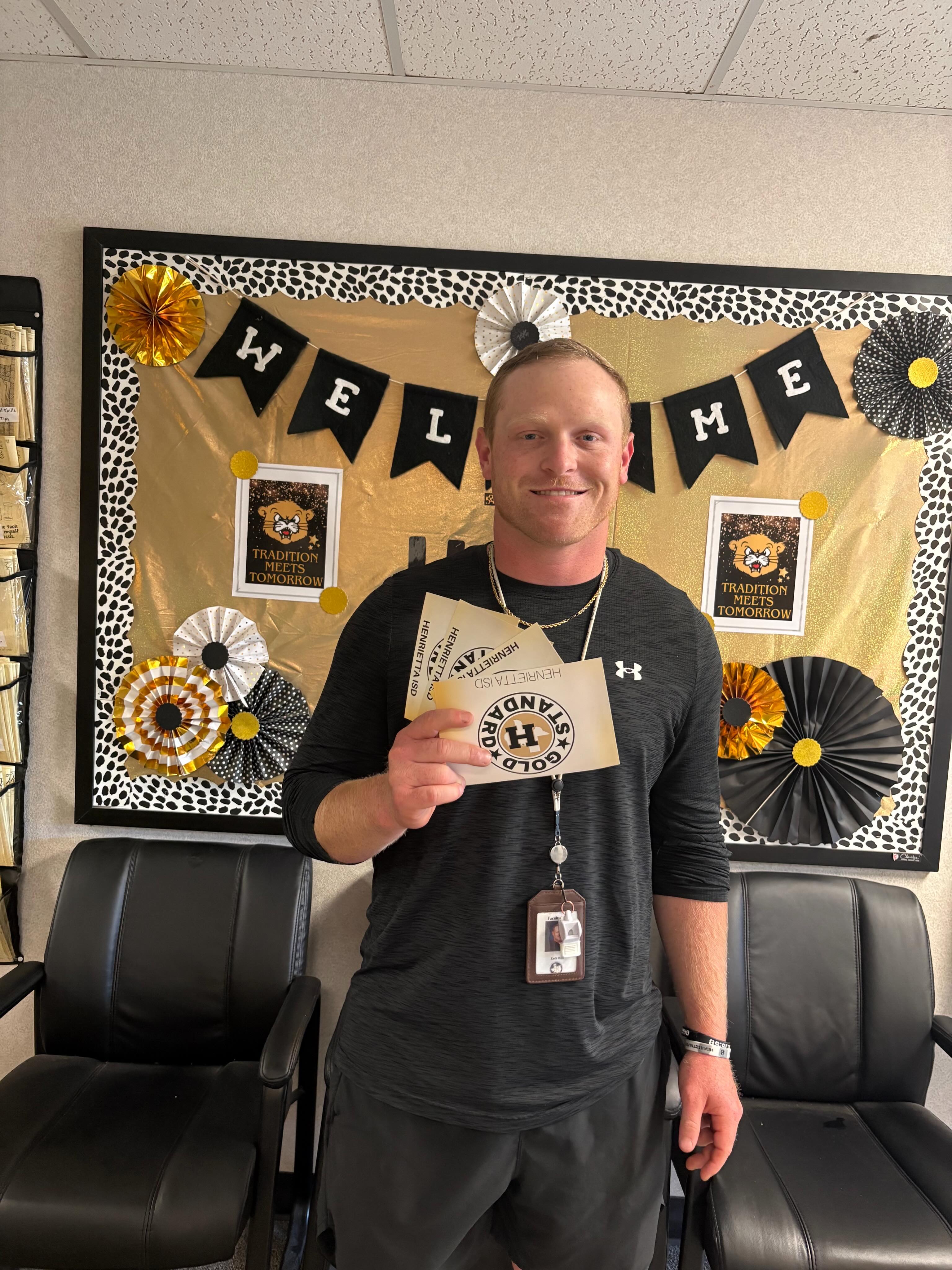 Man in a black shirt holding a Honeybadgers ticket stands in front of a bulletin board with a decorative welcome banner.
