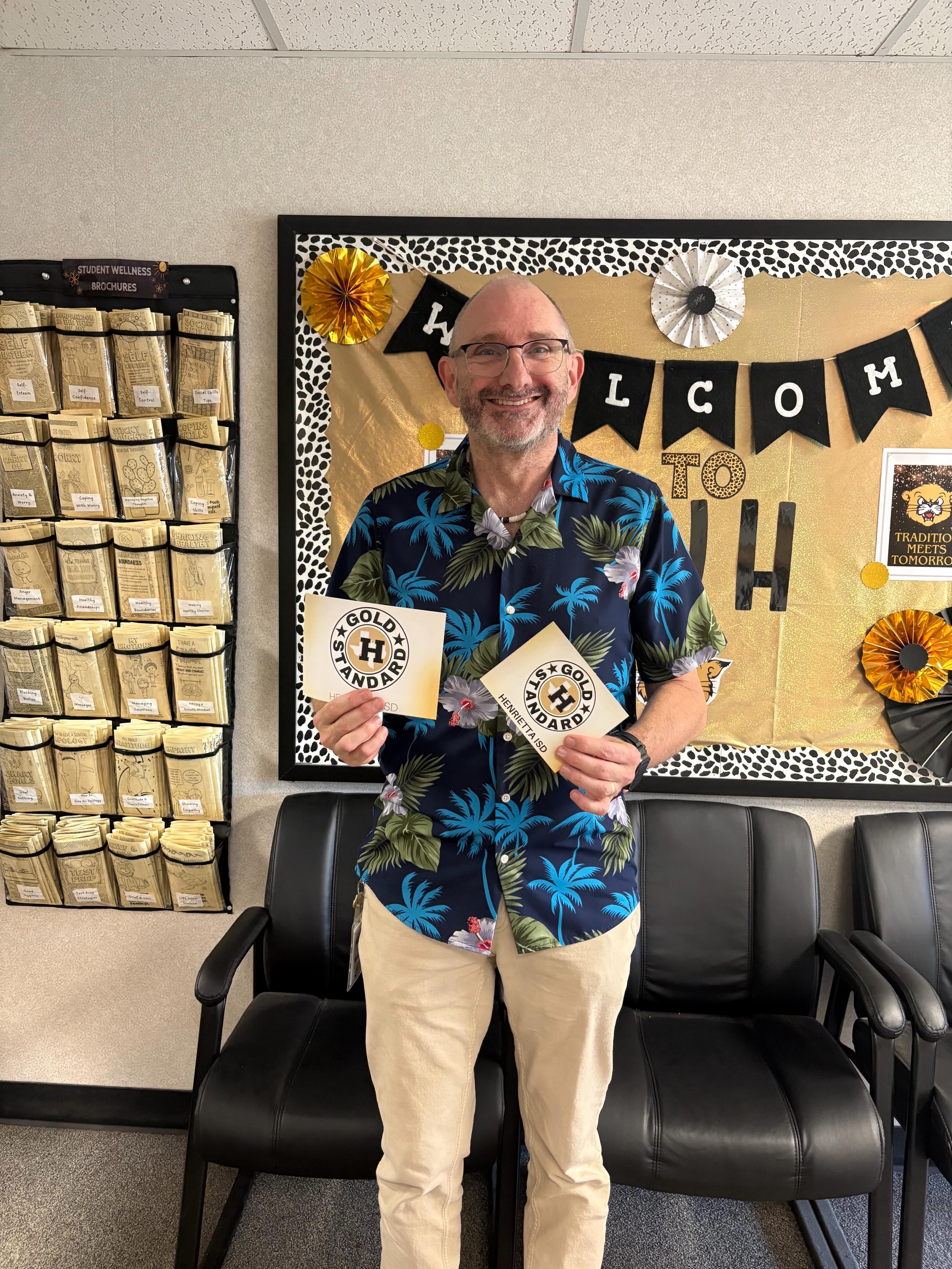 Man stands in front of bulletin board with "Welcome to H" banner, holding two yellow cards.