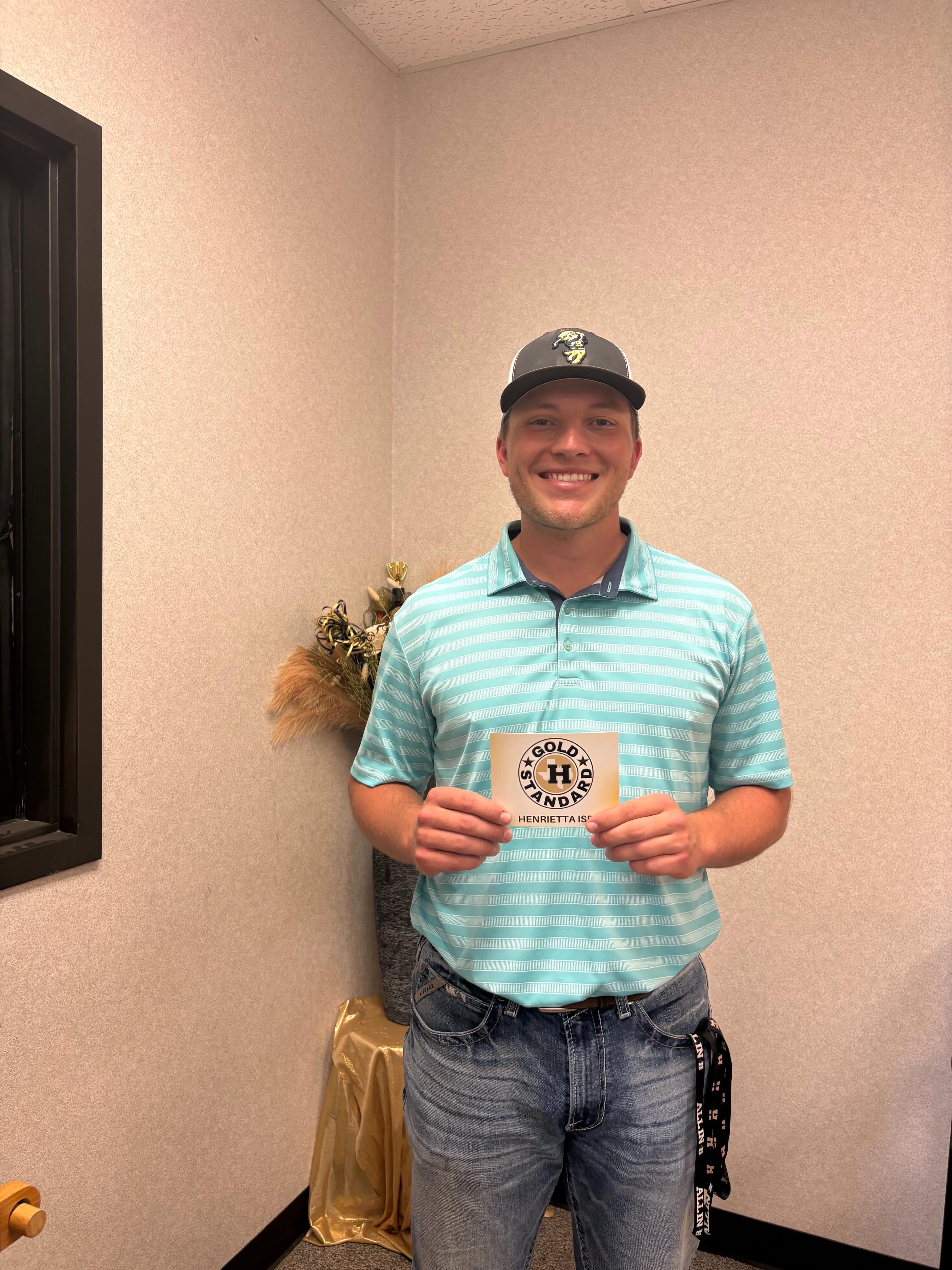 Man in a striped shirt and hat holding a small wooden plaque. He is standing in a room.