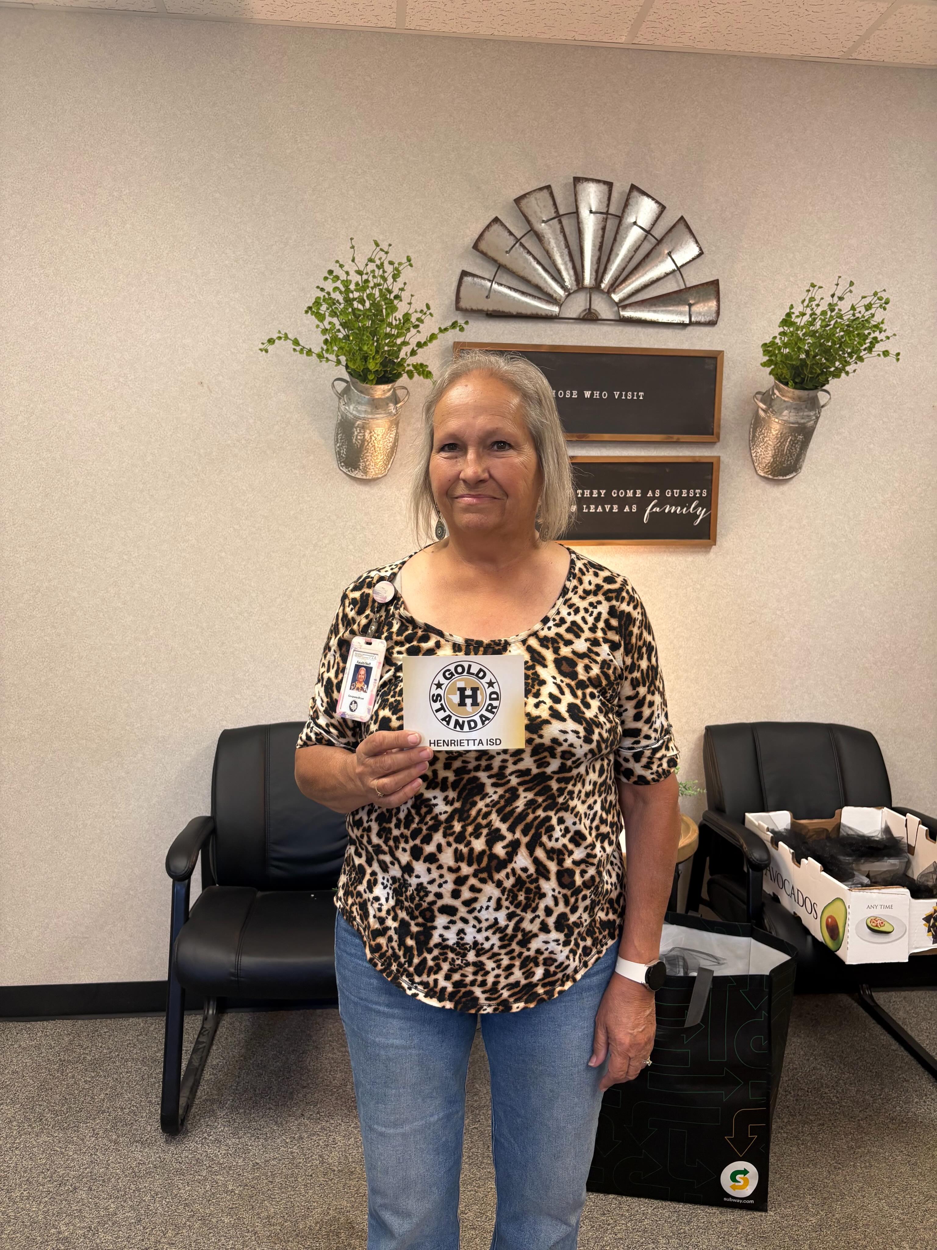 A woman in a leopard print shirt and jeans holds a small card in a room with white walls. Behind her, two potted plants and a decorative wall fan are mounted.